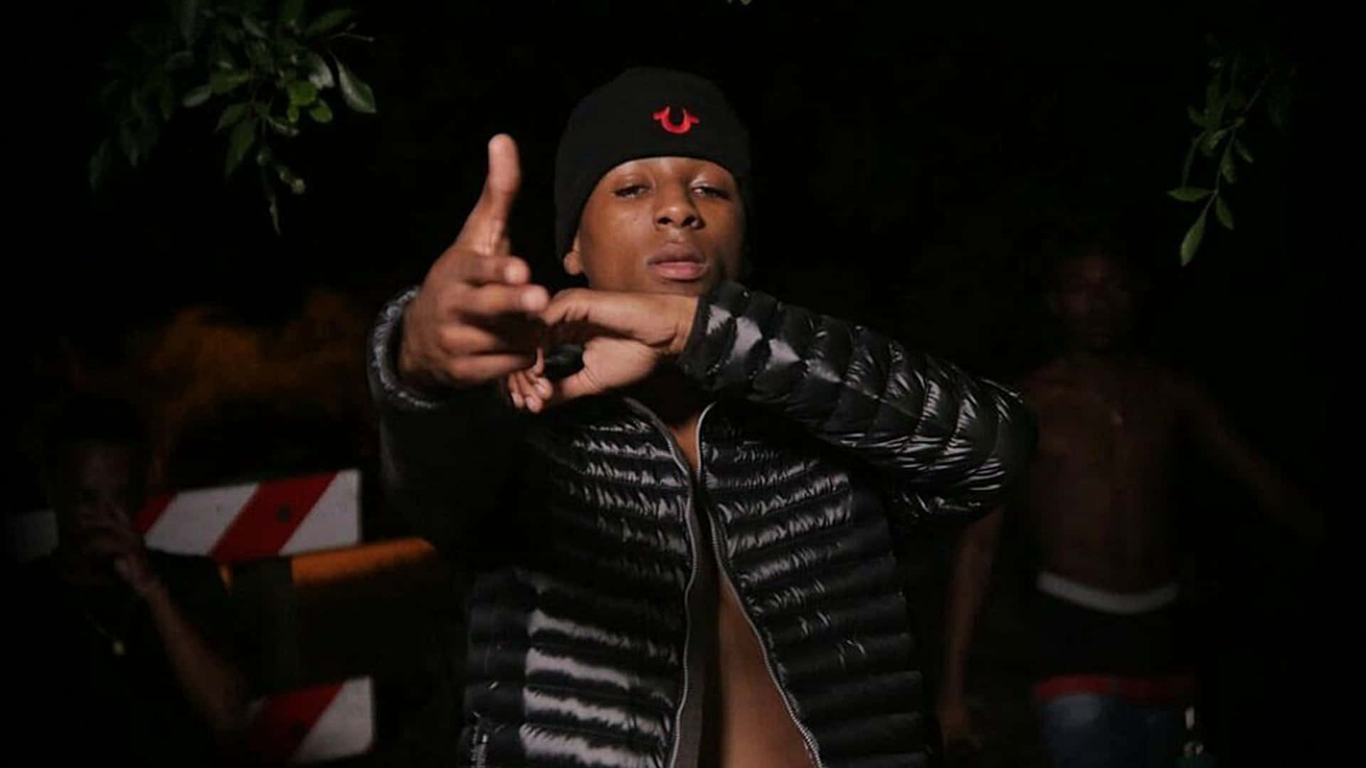 NBA YoungBoy 桌布 壁紙 — 一位身穿黑色羽絨外套和毛帽的年輕男子在昏暗的夜間燈光下，頭頂有樹葉，直接指向相機。