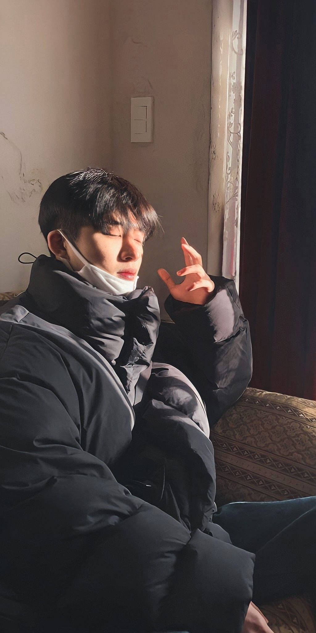 kim hanbin 桌布 壁紙 — 一位身穿黑色羽絨外套的年輕男子坐在窗邊，閉上眼睛，舉起手，沐浴在柔和自然光中。