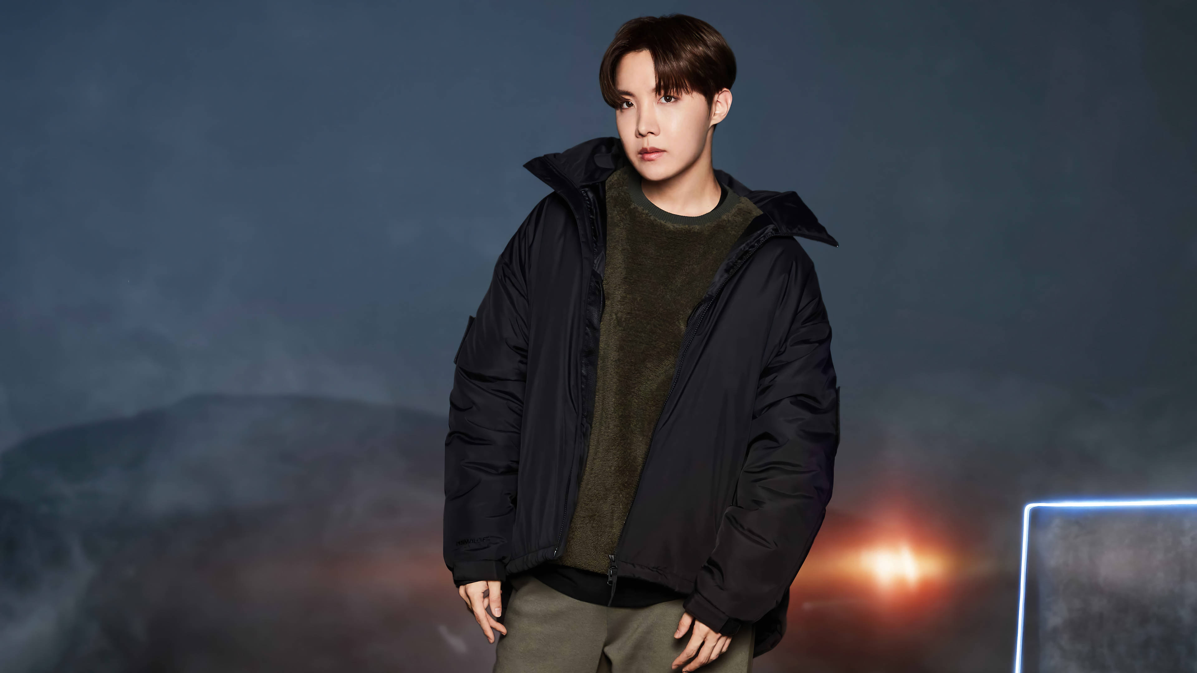j-hope 桌布 壁紙 — 一位身穿黑色羽絨外套的年輕男子自信地站在一個帶有發光橙色燈光和未來感藍色霓虹燈的陰暗背景下。