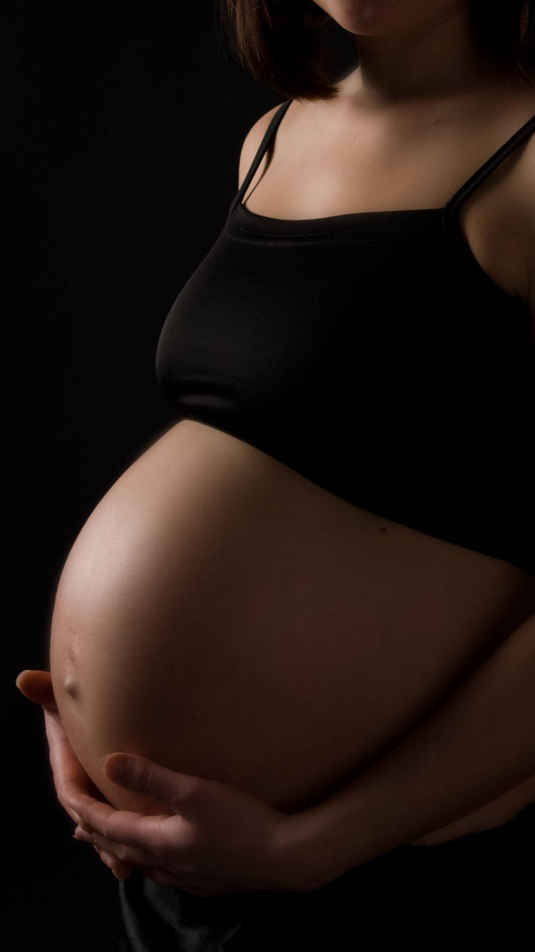 women 桌布 壁紙 — 一位身穿黑色背心的孕婦在柔和燈光下，於深色背景前輕撫腹部。