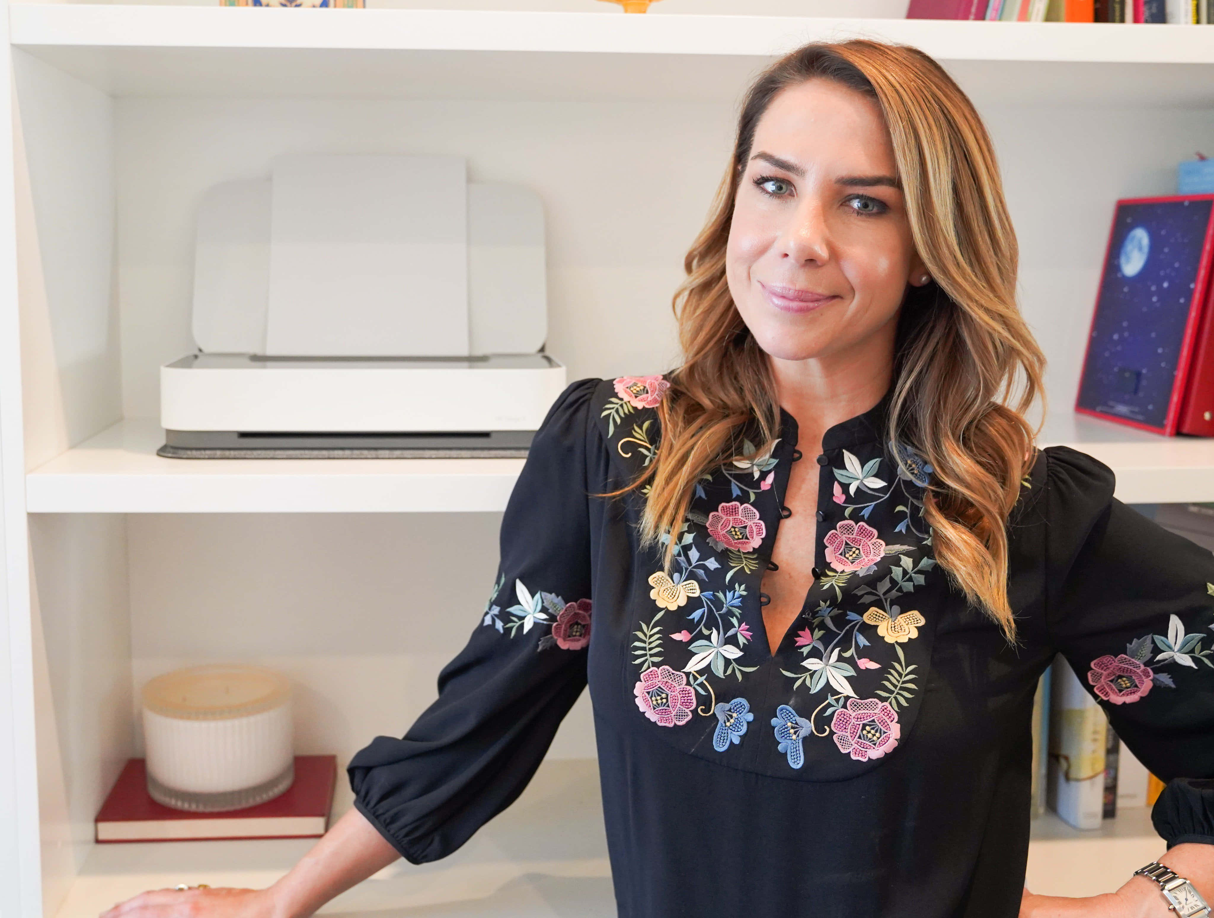 Kate Ritchie 桌布 壁紙 — 一位身穿黑色花卉刺繡襯衫的女性微笑著靠在一個放有印表機和蠟燭的白色書架上。