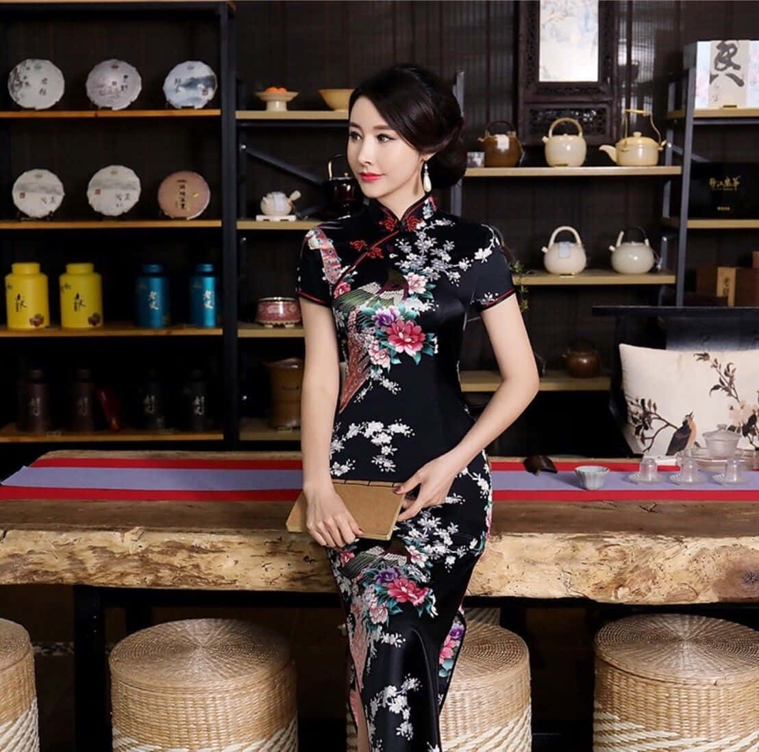 Long Dress 桌布 壁紙 — 一位身穿黑色花卉旗袍的女性，優雅地站在傳統茶莊的質樸木桌旁，店內擺滿陶瓷器皿。