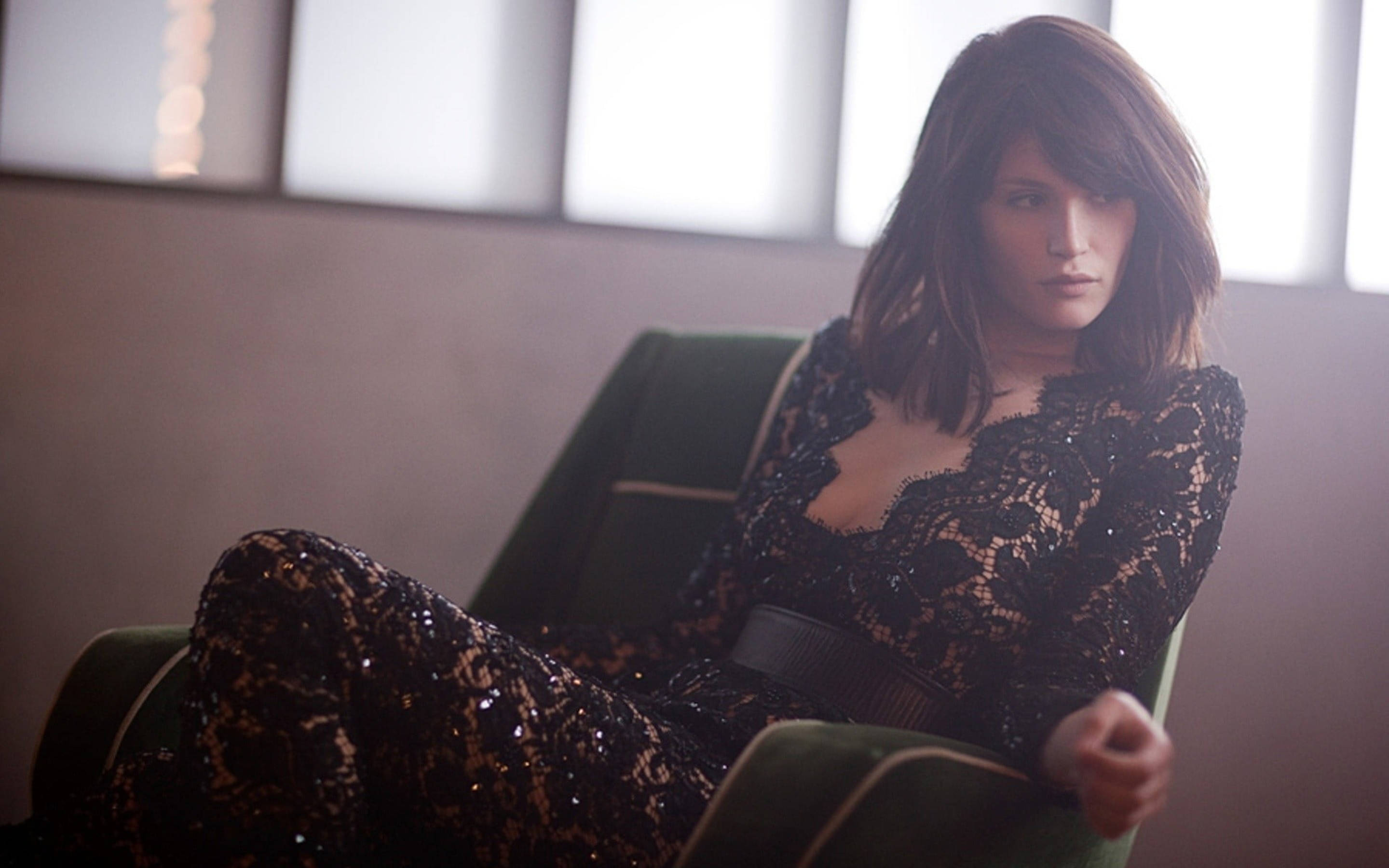 Gemma Arterton 桌布 壁紙 — 一位身穿黑色蕾絲服裝的女性，端坐在綠色天鵝絨椅子上，柔和的光線從她身後的窗戶透進來。