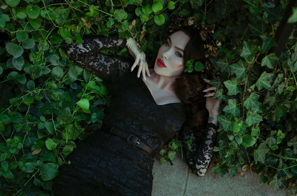 Gothic Fashion 桌布 壁紙 — 一位身穿黑色蕾絲裙的女性躺在茂密的綠色常春藤中，擁有鮮豔的紅唇和白色指甲。