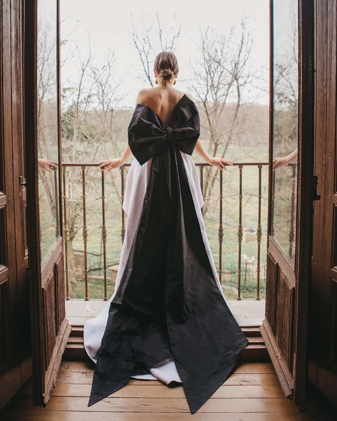 Black And White Wedding 桌布 壁紙 — 一位身穿黑色蝴蝶結禮服的女士站在木製陽台上，俯瞰著陰天下的光禿樹木和田野。