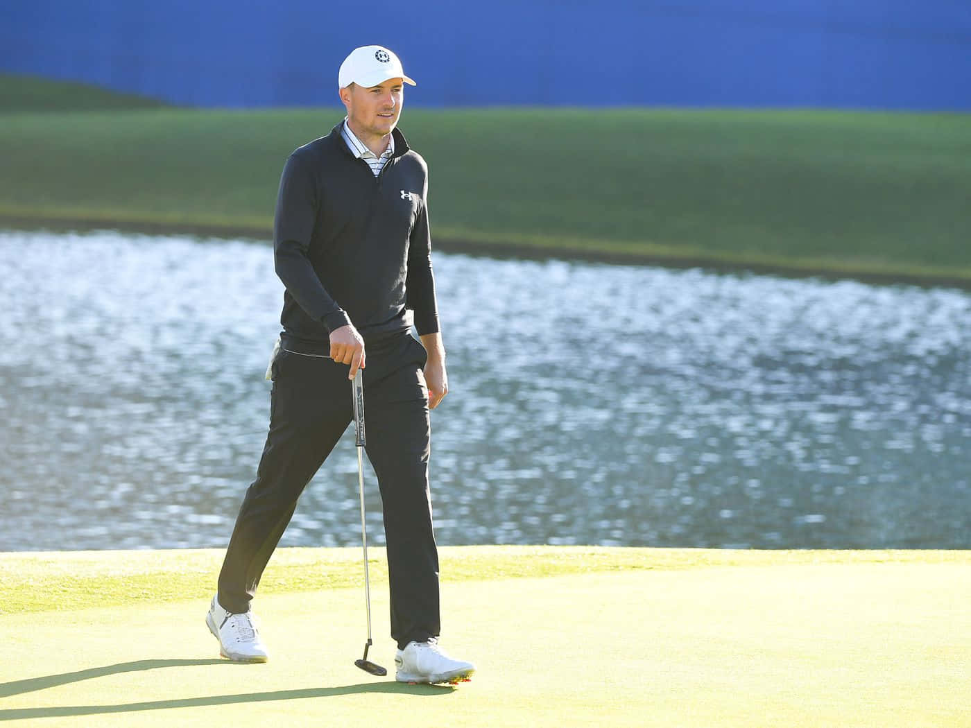 Golfing 桌布 壁紙 — 一位身穿黑色衣服、戴白色帽子的高爾夫球手在陽光明媚的果嶺上行走，身後有水障礙。