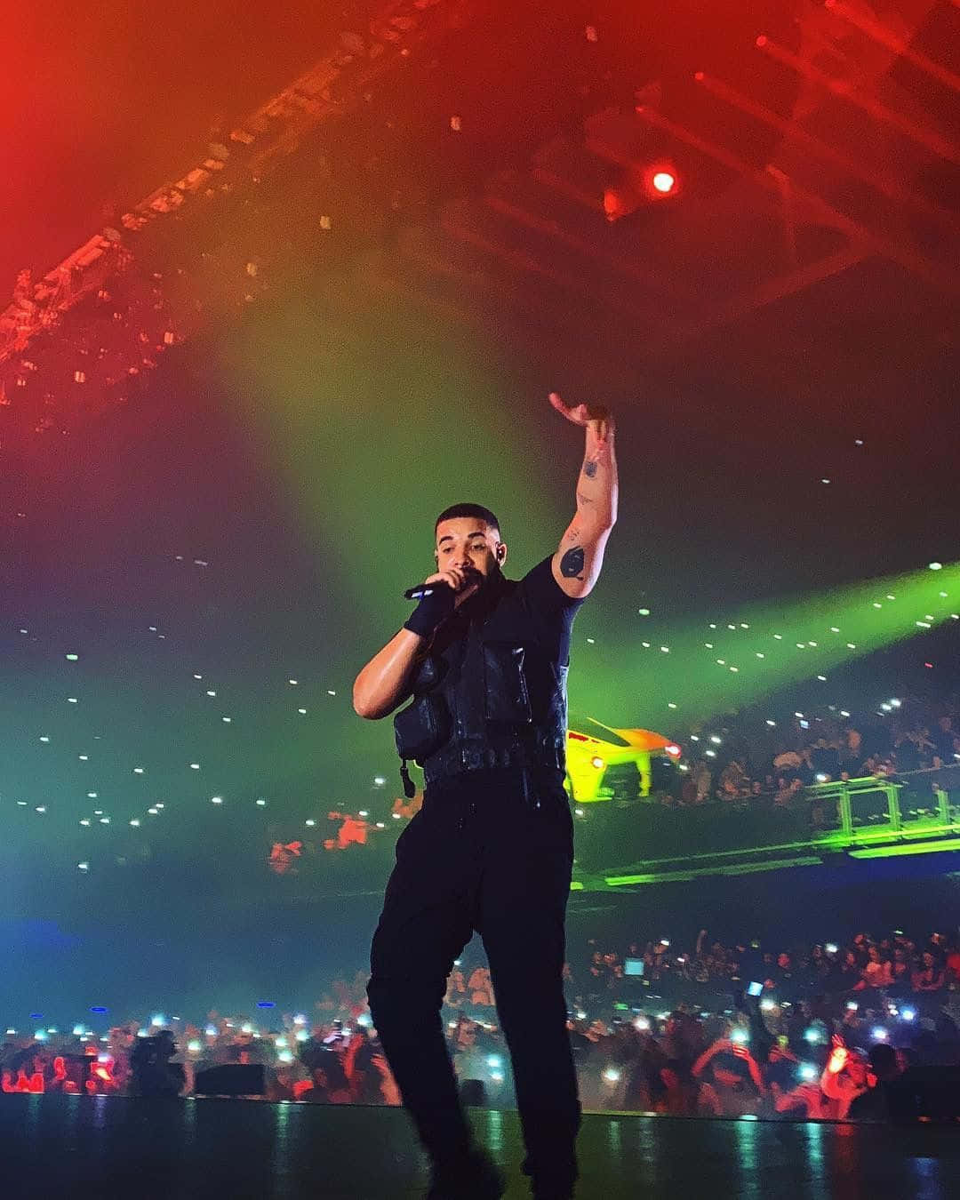 Drake 桌布 壁紙 — 一位身穿黑色衣服的男性表演者在燈光璀璨的舞台上，對著麥克風唱歌，舞台上有紅色和綠色的聚光燈，觀眾舉起手機。