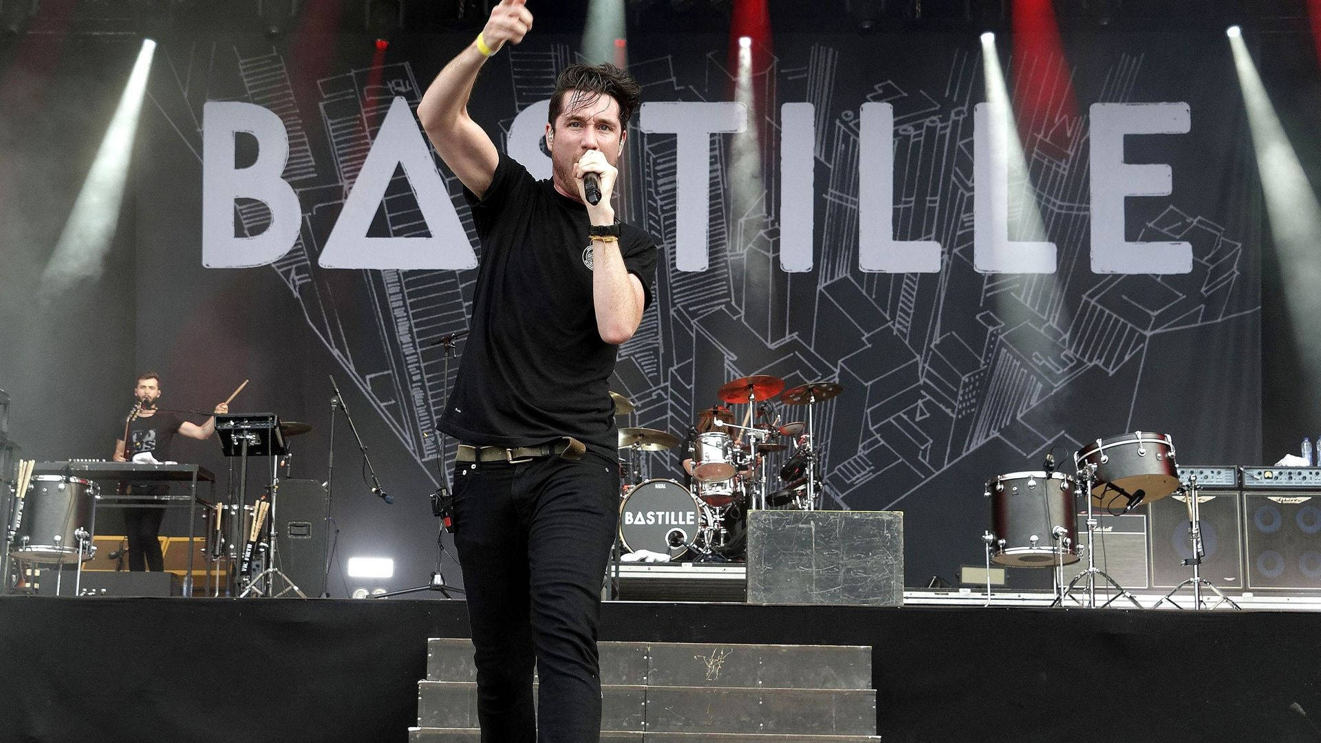 Bastille 桌布 壁紙 — 一位身穿黑色衣服的男歌手在明亮的聚光燈下與樂隊在'BASTILLE'背景前充滿活力地表演。