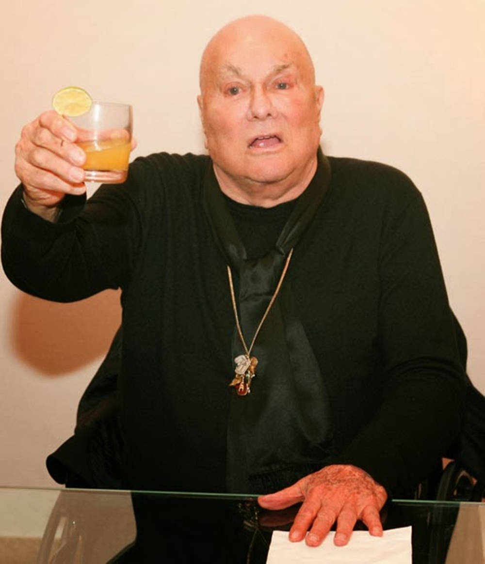 Celebrity 桌布 壁紙 — 一位身穿黑色衣服的禿頭長者，坐在玻璃桌前，舉起一杯帶有萊姆裝飾的橙色飲品。