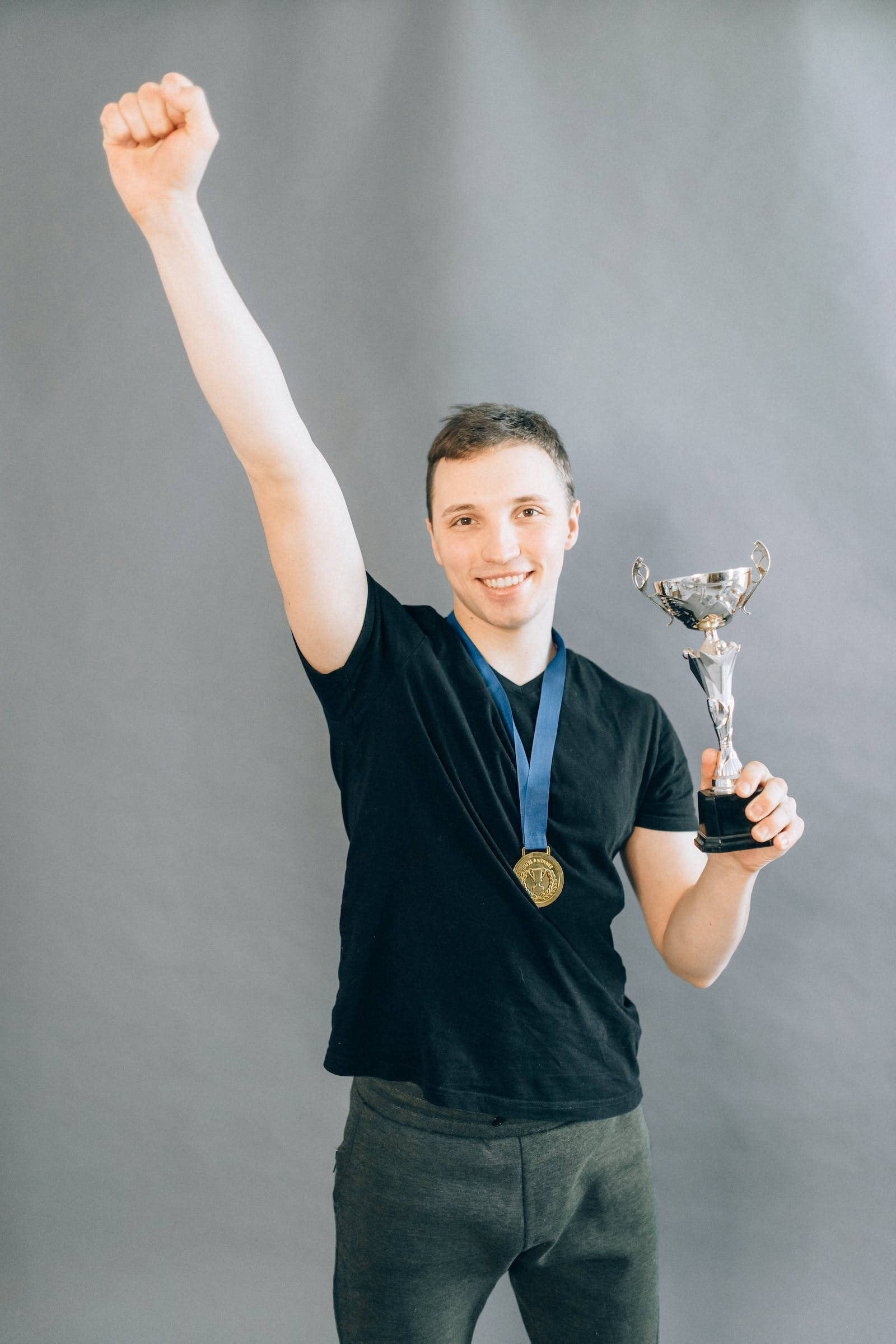 youre the best 桌布 壁紙 — 一位身穿黑色襯衫的勝利年輕人，自豪地高舉銀色獎盃並佩戴金牌，同時握緊拳頭。