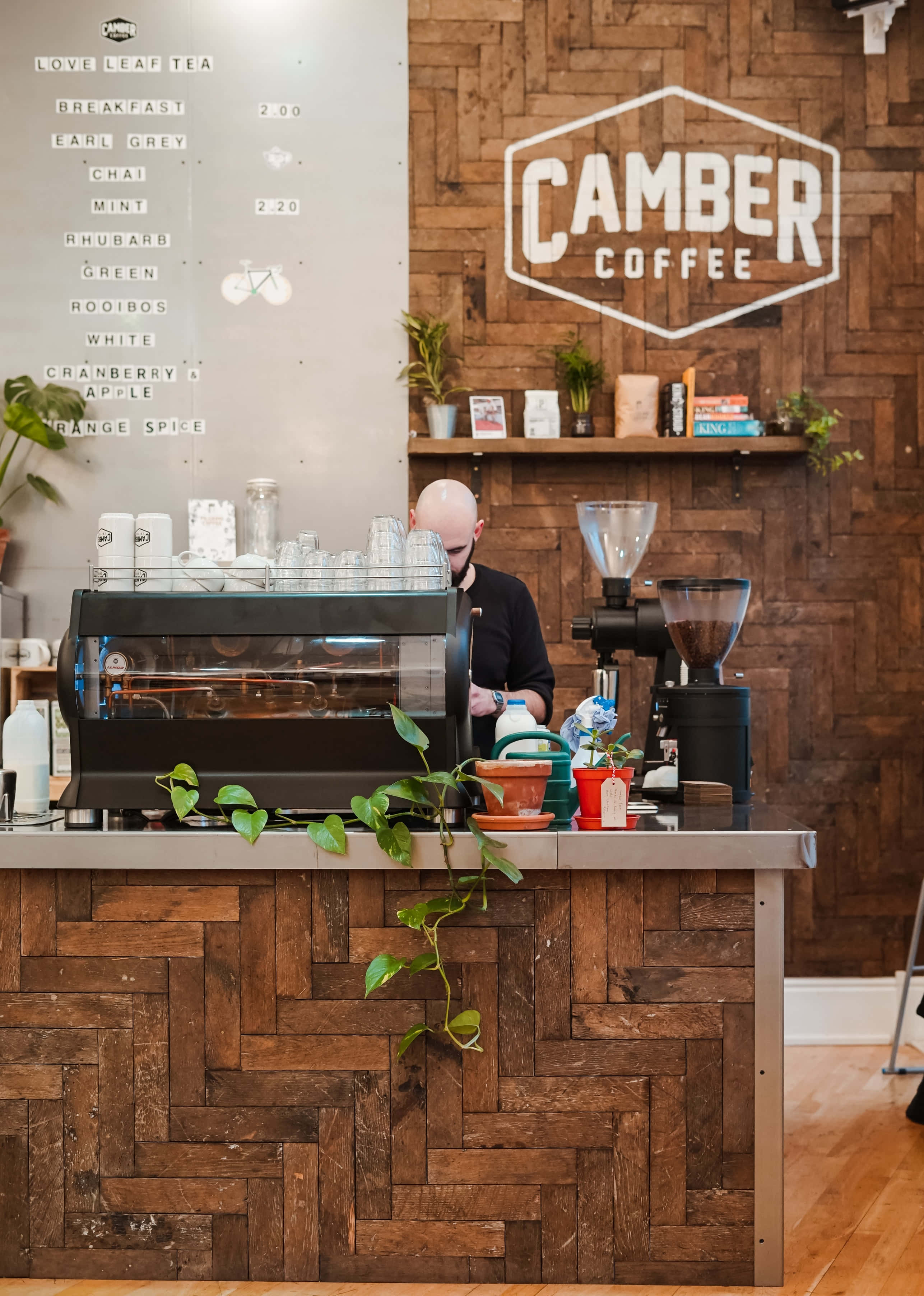 Coffee Aesthetic 桌布 壁紙 — 一位身穿黑色襯衫的咖啡師在一個舒適的咖啡館裡，在木製人字形櫃檯後面準備咖啡，店內有 'Camber Coffee' 標誌。
