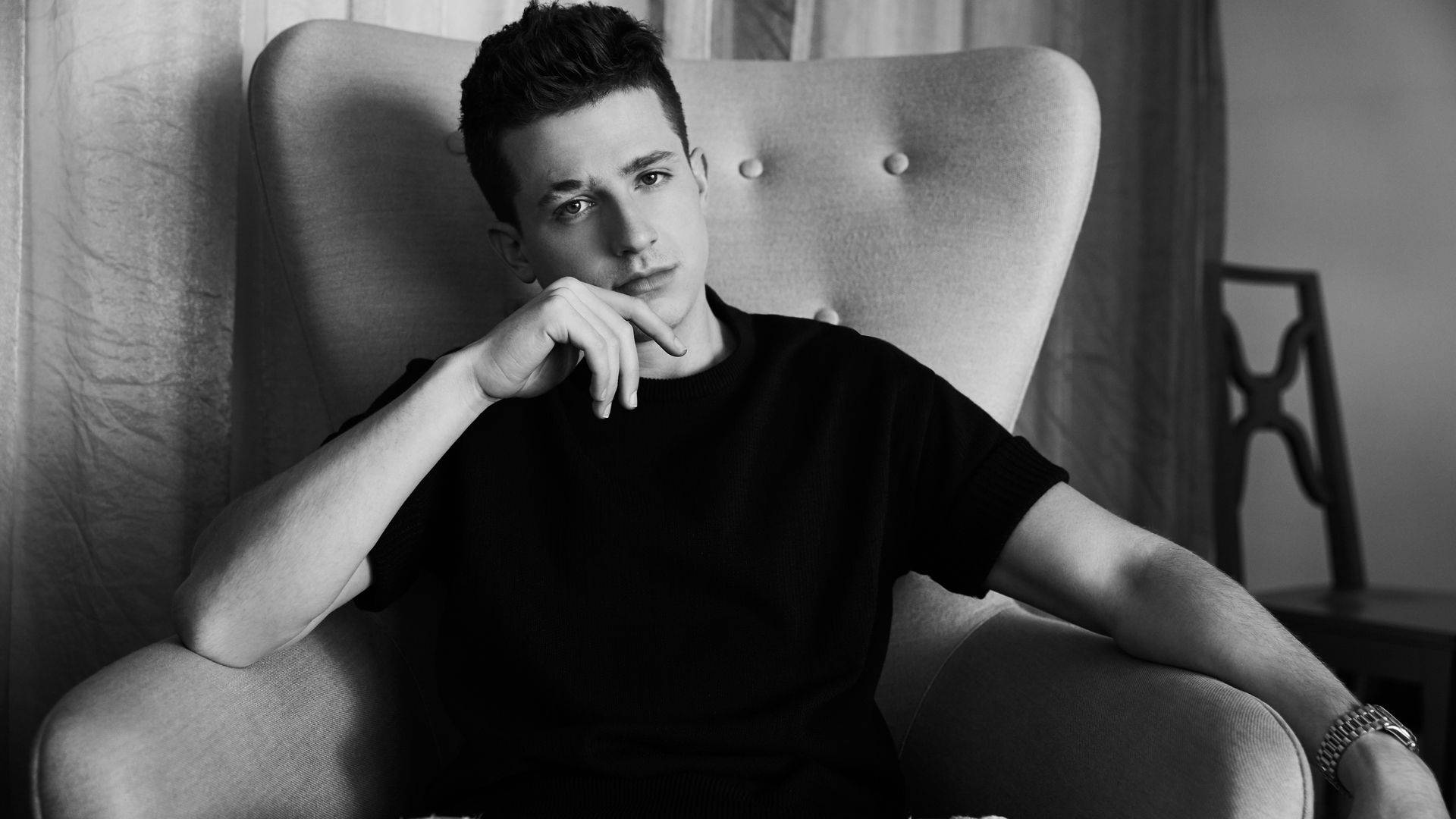 Charlie Puth 桌布 壁紙 — 一位身穿黑色襯衫的年輕男子若有所思地坐在簇絨扶手椅上，手托下巴，一幅單色室內肖像。