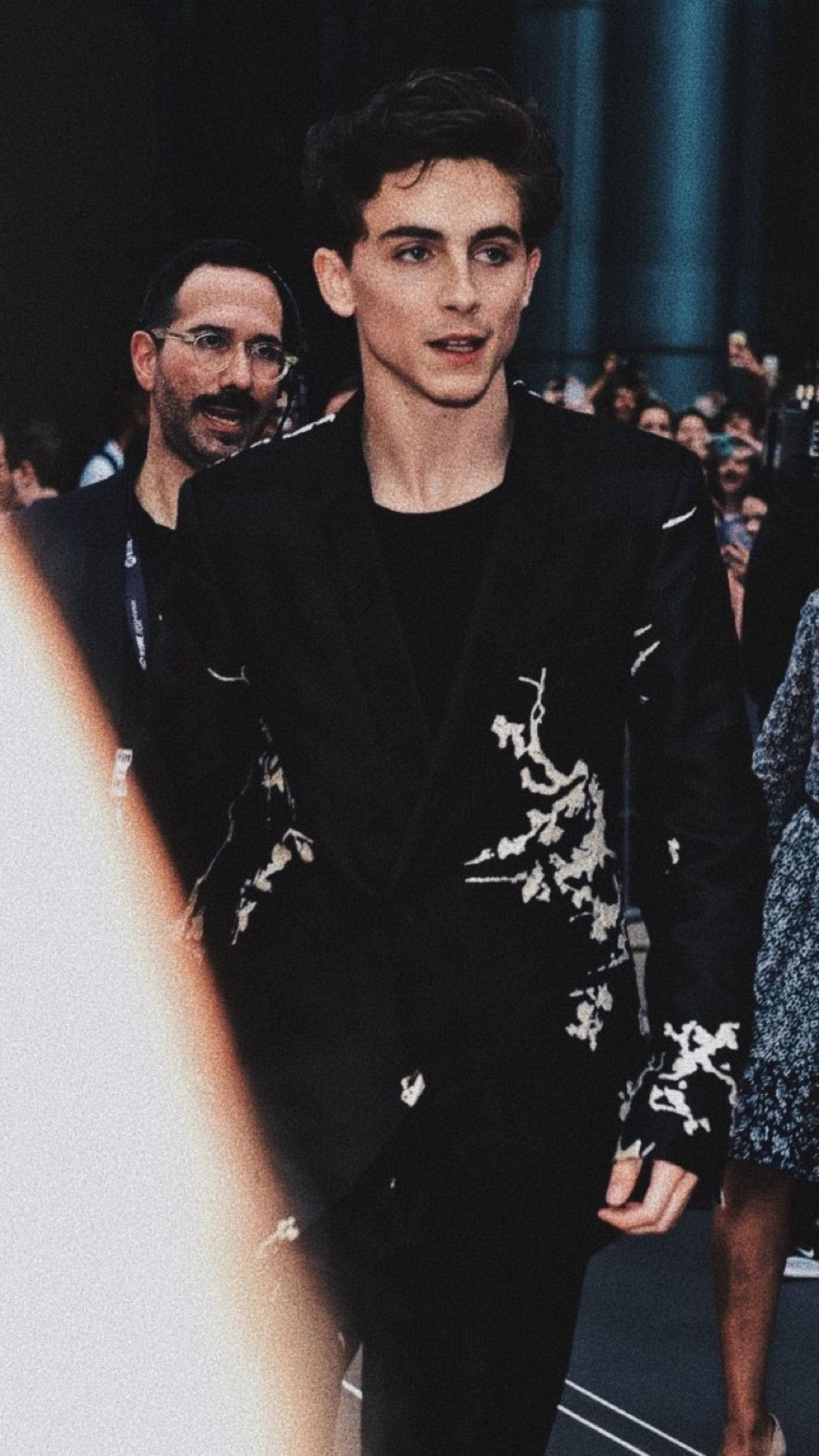 Timothée Chalamet 桌布 壁紙 — 一位身穿黑色西裝、帶有白色花卉刺繡的年輕男子自信地走在時裝伸展台上，背景有觀眾。