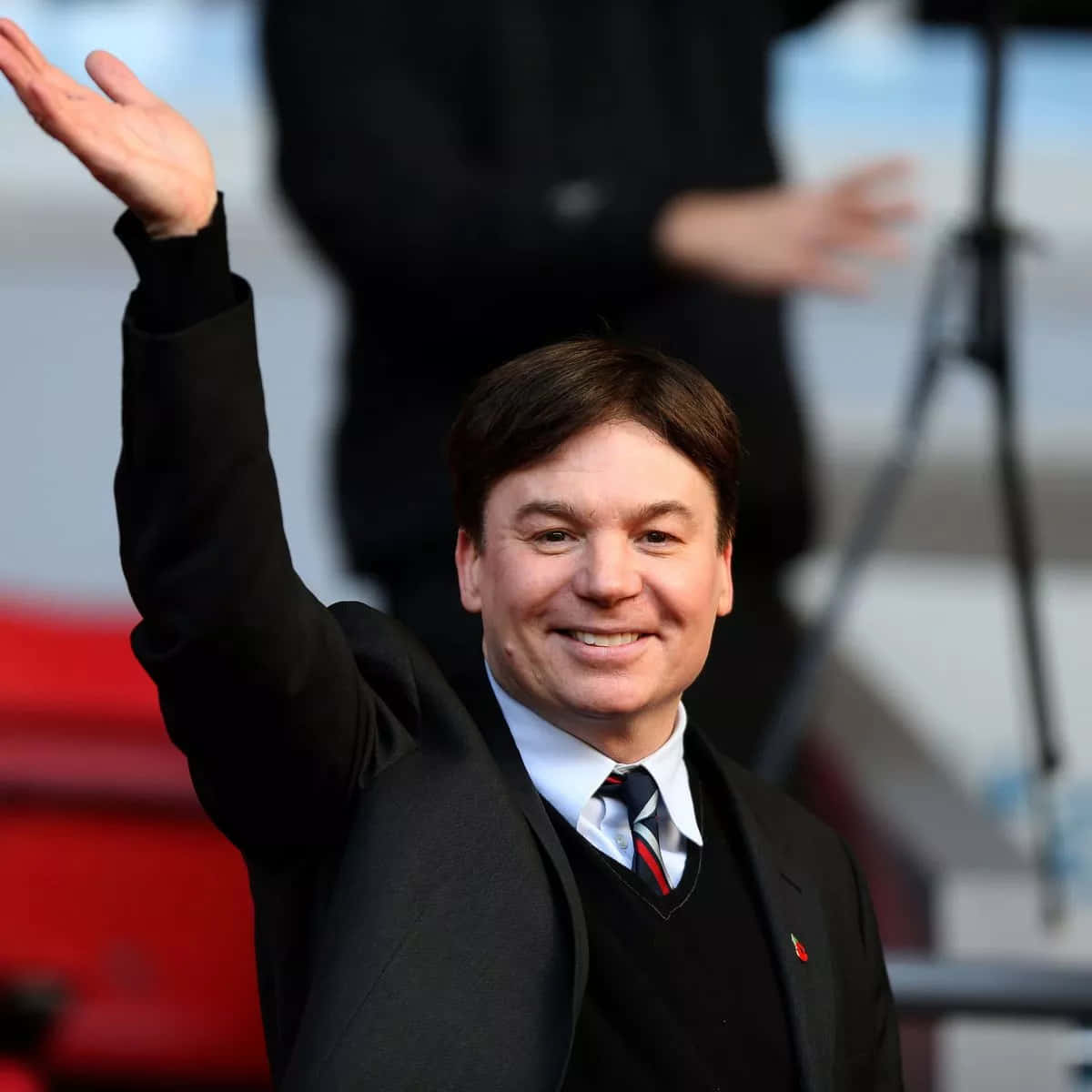Mike Myers 桌布 壁紙 — 一位身穿黑色西裝、面帶微笑的男子，在戶外活動中熱情揮手，背景有紅地毯和模糊的人物。
