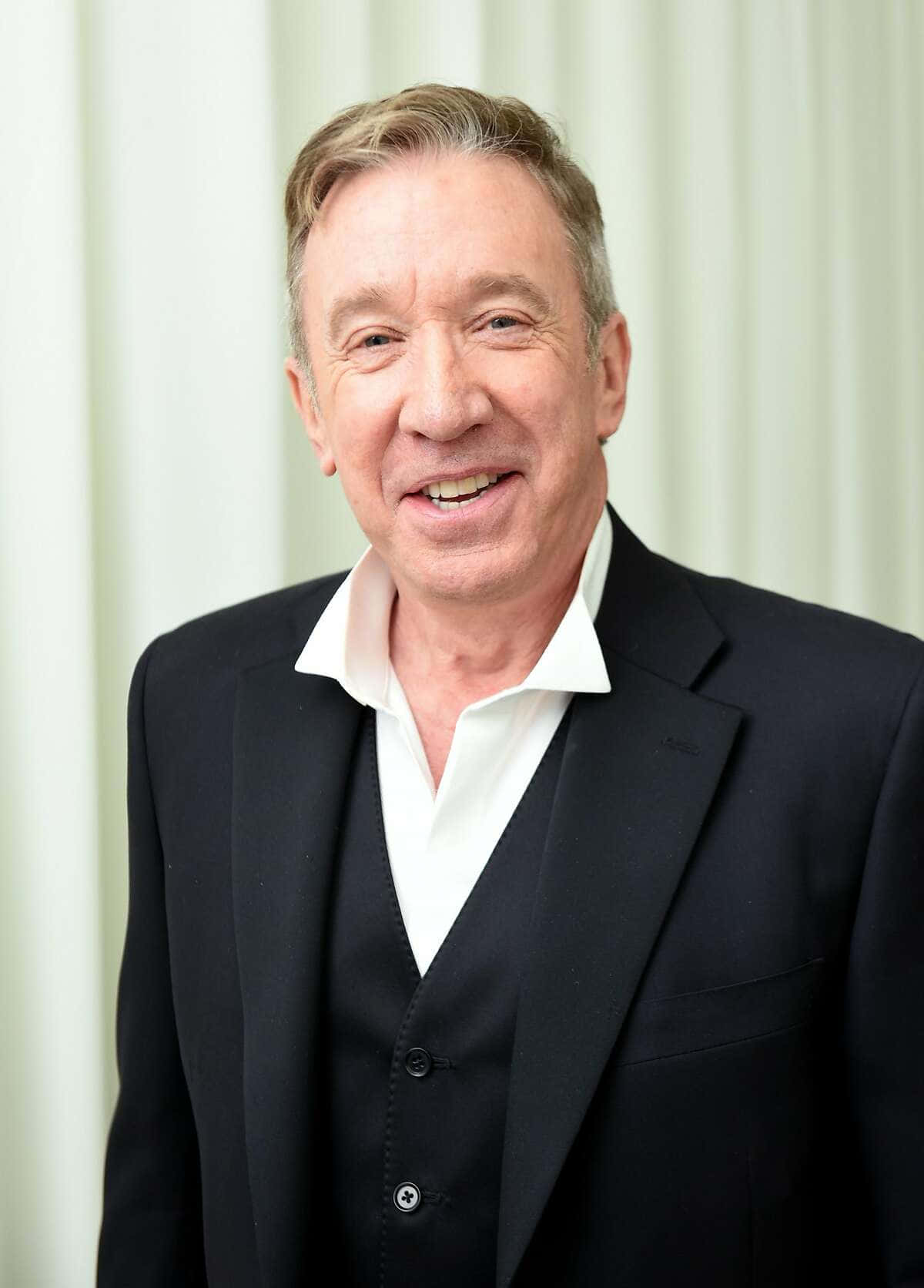 Tim Allen 桌布 壁紙 — 一位身穿黑色西裝和白色襯衫的開朗中年男子，在柔和的奶油色窗簾背景下溫暖地微笑。