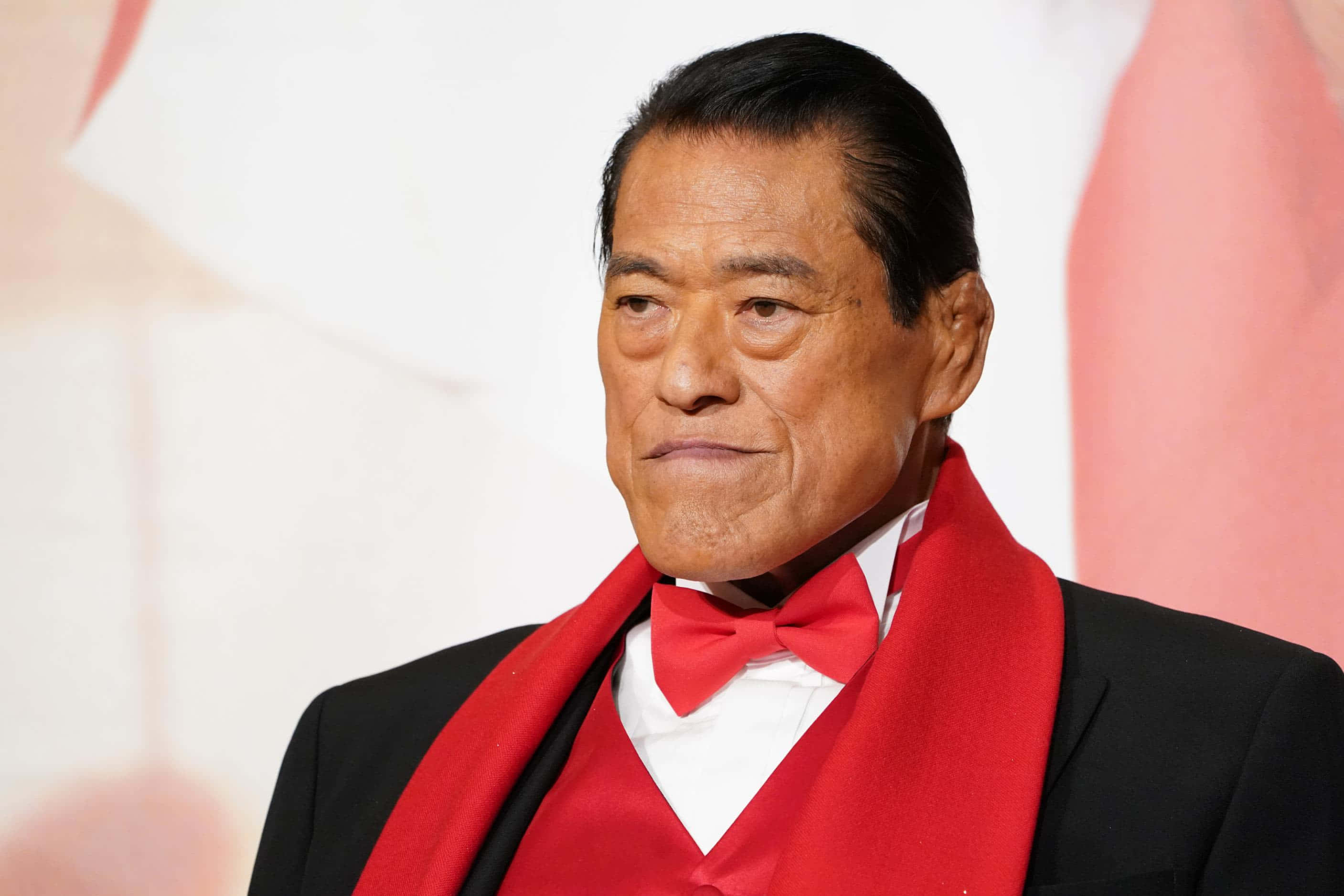 運動 桌布 壁紙 — 一位身穿黑色西裝和紅色領結的尊貴男士，在柔和的粉紅色和白色背景前，表情嚴肅地站立。