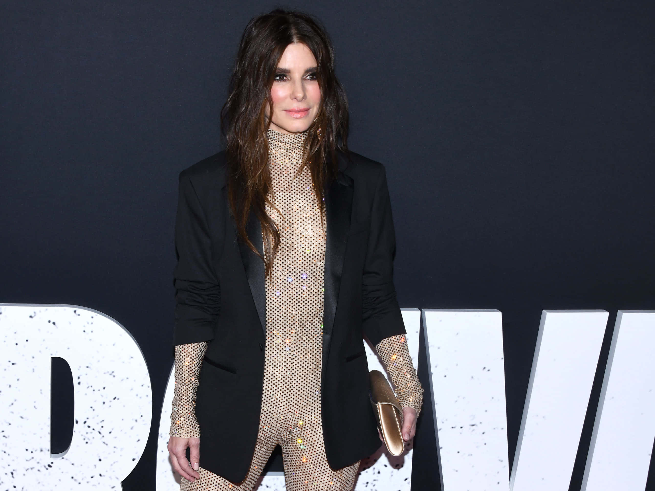 Sandra Bullock 2021 桌布 壁紙 — 一位身穿黑色西裝外套、內搭閃亮金色連身衣的女性，在深色背景前自信地擺姿勢，背景上有大型白色字母。