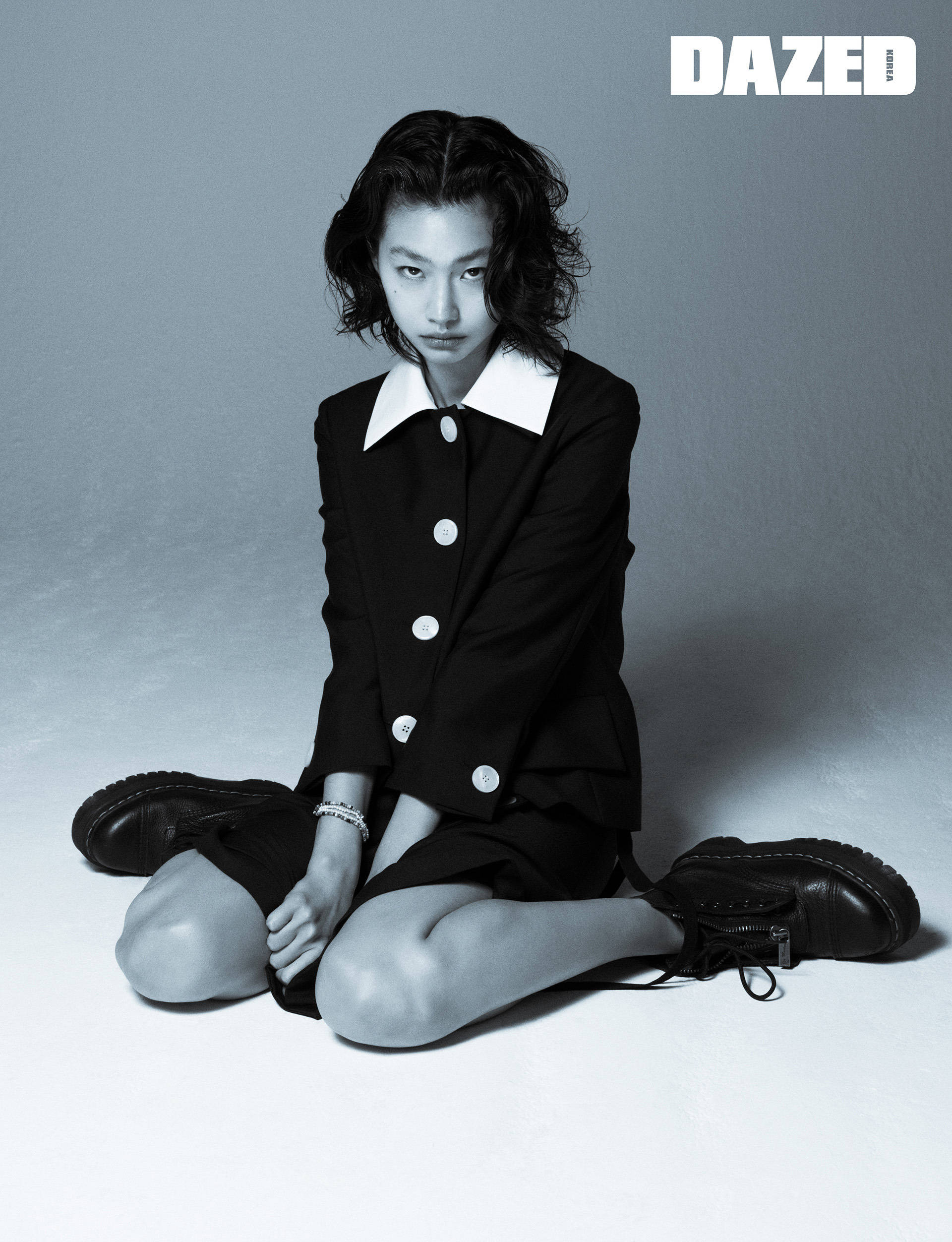 Hoyeon 桌布 壁紙 — 一位身穿黑色西裝外套、白色領口的年輕女性，在素色地板上盤腿而坐，於單色調工作室拍攝中凝視鏡頭。