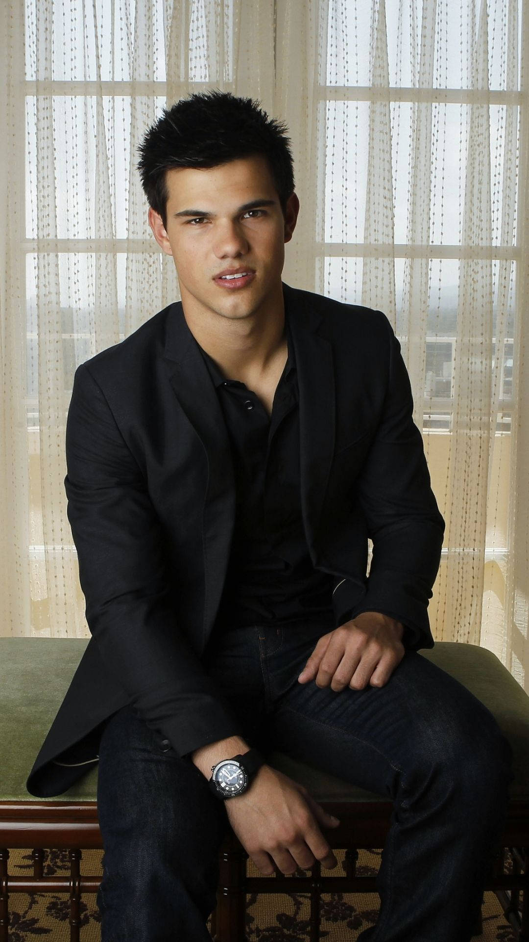 Taylor Lautner 桌布 壁紙 — 一位身穿黑色西裝外套和牛仔褲的年輕男子自信地坐在有圖案的長椅上，前面是薄紗窗簾。
