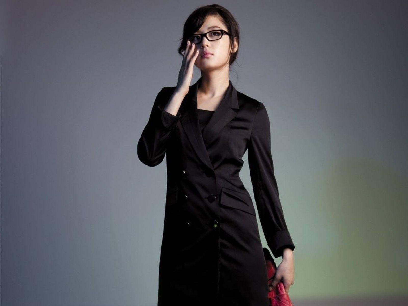 Jun Ji Hyun 桌布 壁紙 — 一位身穿黑色西裝外套和眼鏡的優雅女性，在柔和的灰色漸變背景上輕輕觸碰臉龐。