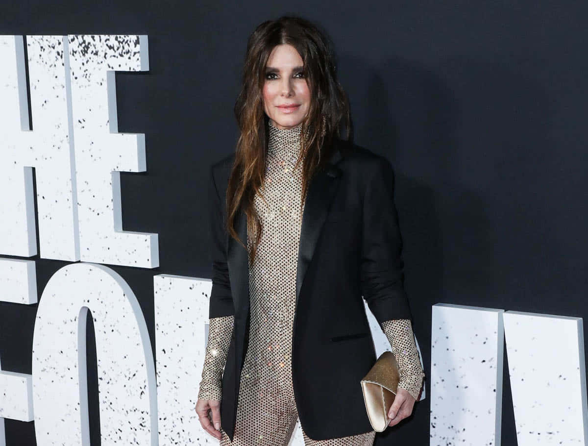 Sandra Bullock 2021 桌布 壁紙 — 一位身穿黑色西裝外套和閃亮金色連衣裙的女性在紅地毯活動中自信地擺姿勢，身後有大型白色字母。