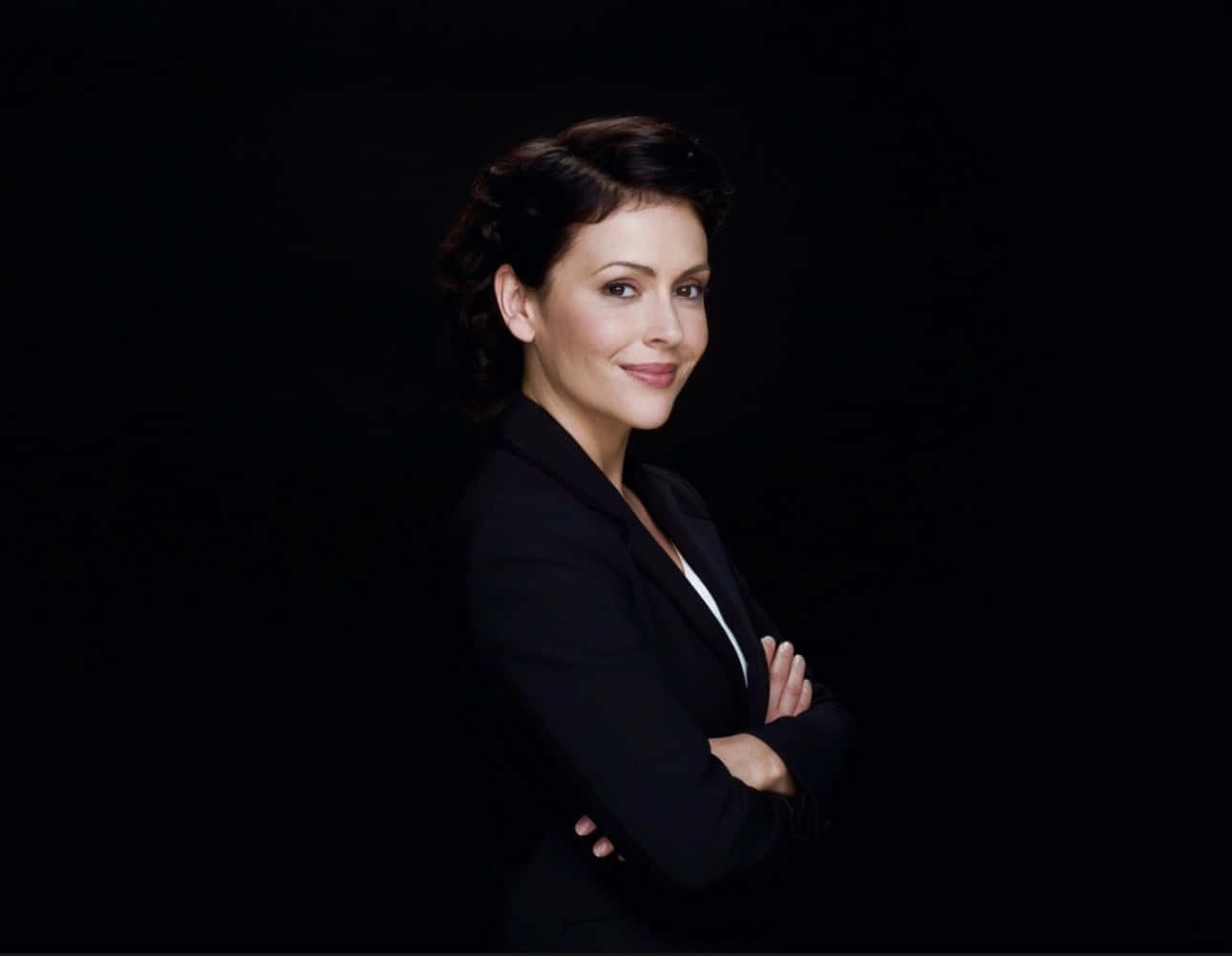 Charmed Season 7 Promotional Image - Alyssa Milano 壁紙 — 一位身穿黑色西裝外套的女性自信地交叉雙臂，站在純黑色背景前，對著鏡頭微笑。