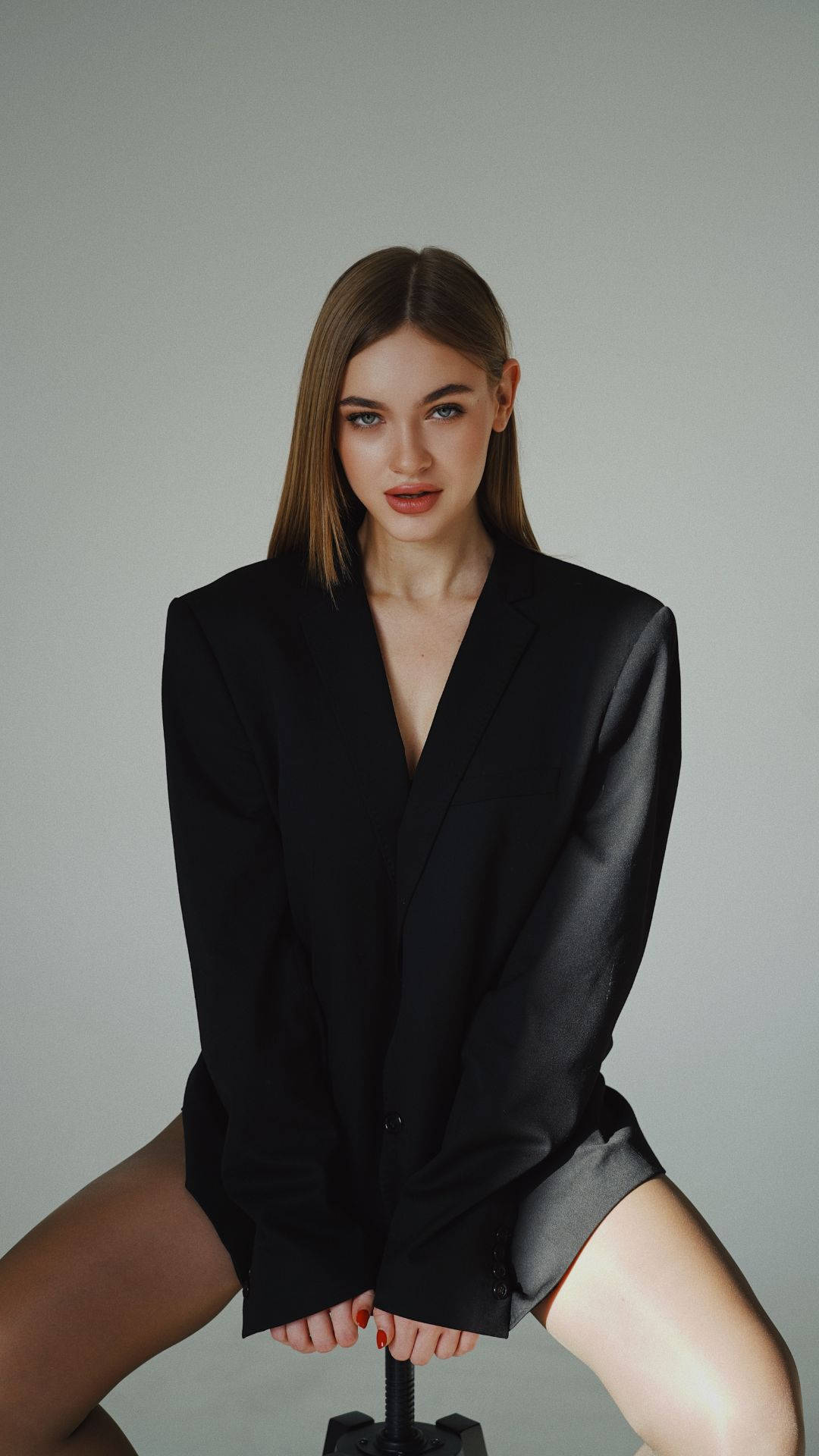 suit 桌布 壁紙 — 一位身穿黑色西裝外套的女性自信地坐在灰色背景前的凳子上，直視鏡頭。