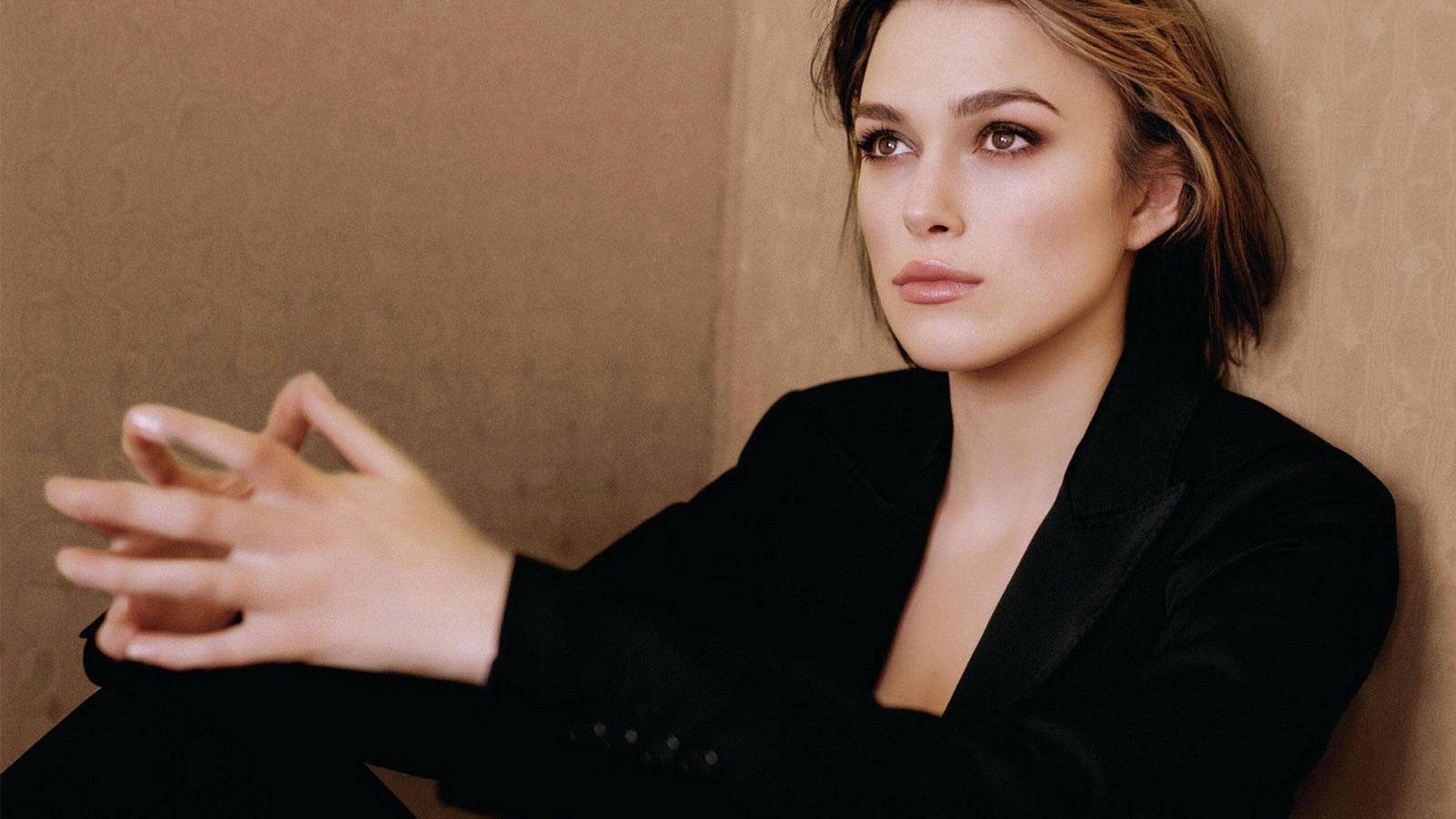 Keira Knightley 桌布 壁紙 — 一位身穿黑色西裝外套的女性靠著帶有紋理的米色牆壁，眼神若有所思，雙手輕輕交握。