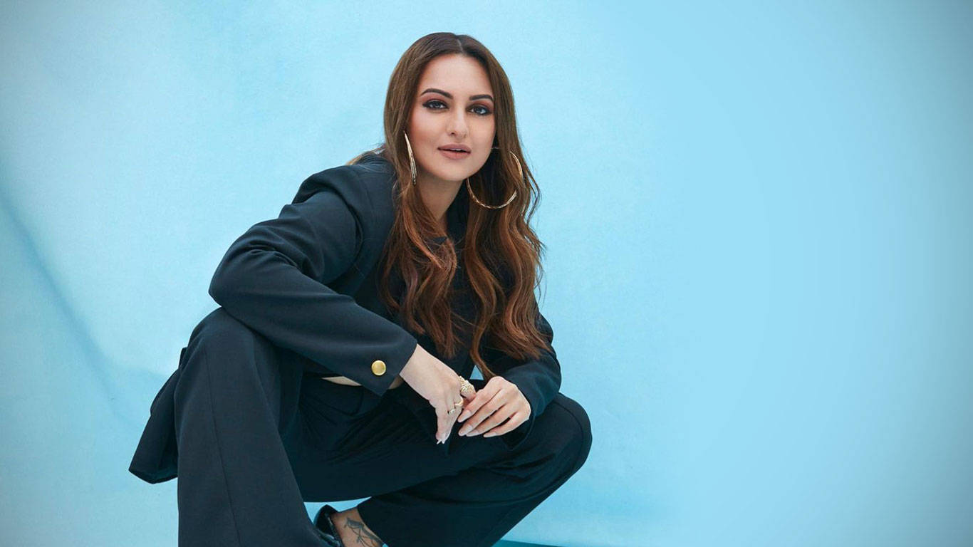 Sonakshi Sinha 桌布 壁紙 — 一位身穿黑色西裝的女性自信地蹲在柔和的藍色攝影棚背景前，長髮波浪，戴著金色圓環耳環。