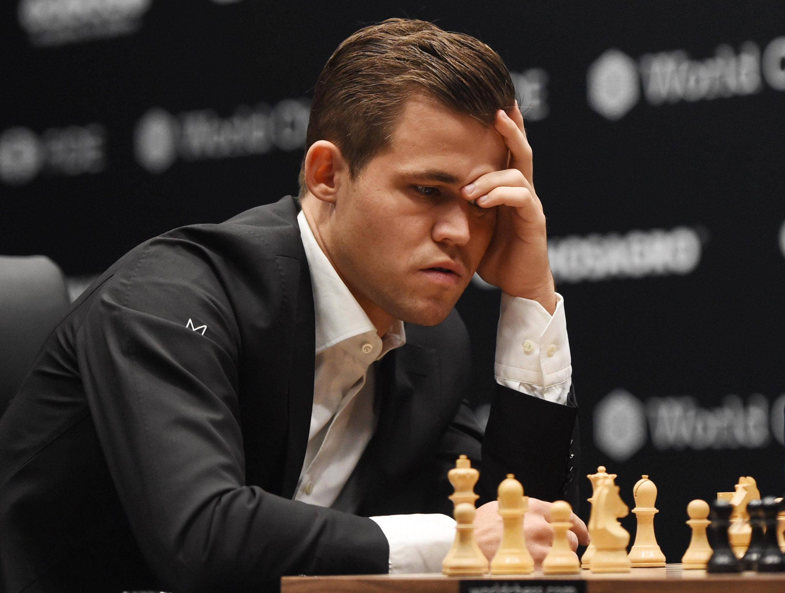 Magnus 桌布 壁紙 — 一位身穿黑色西裝的專注棋手，將頭靠在手上，面前棋盤上的棋子正在對弈中。