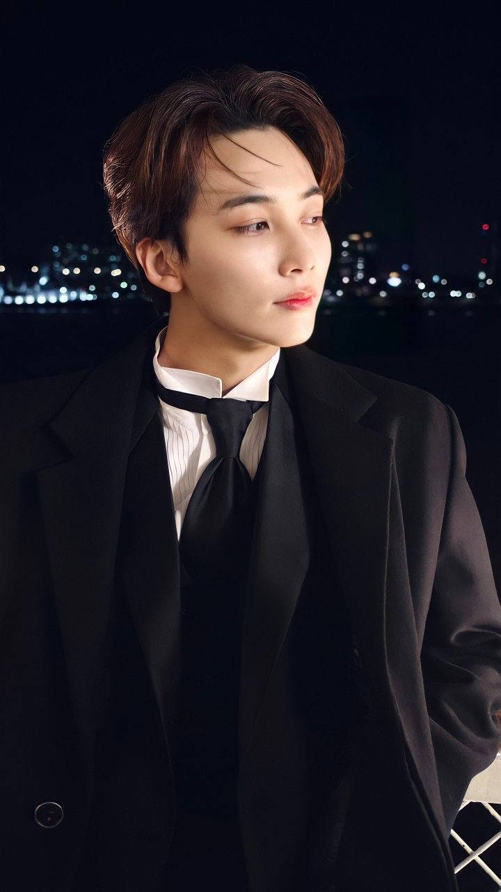 yoon jeonghan 桌布 壁紙 — 一位身穿黑色西裝的年輕男子，在模糊的城市夜景和閃爍燈光中若有所思地凝視。