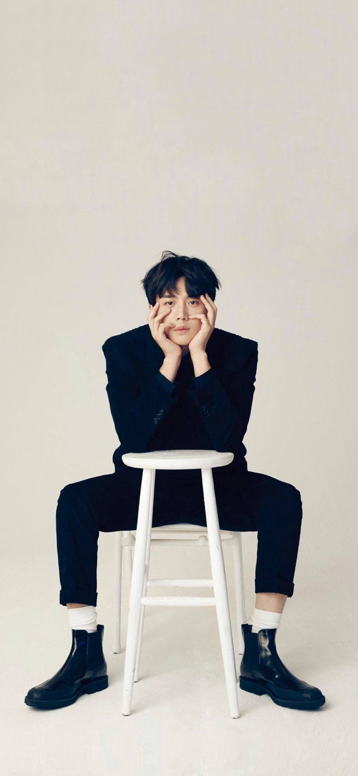 Kim Seon Ho 桌布 壁紙 — 一位身穿黑色西裝的年輕男子坐在白色凳子上，雙手框著臉，背景是純色淺色。
