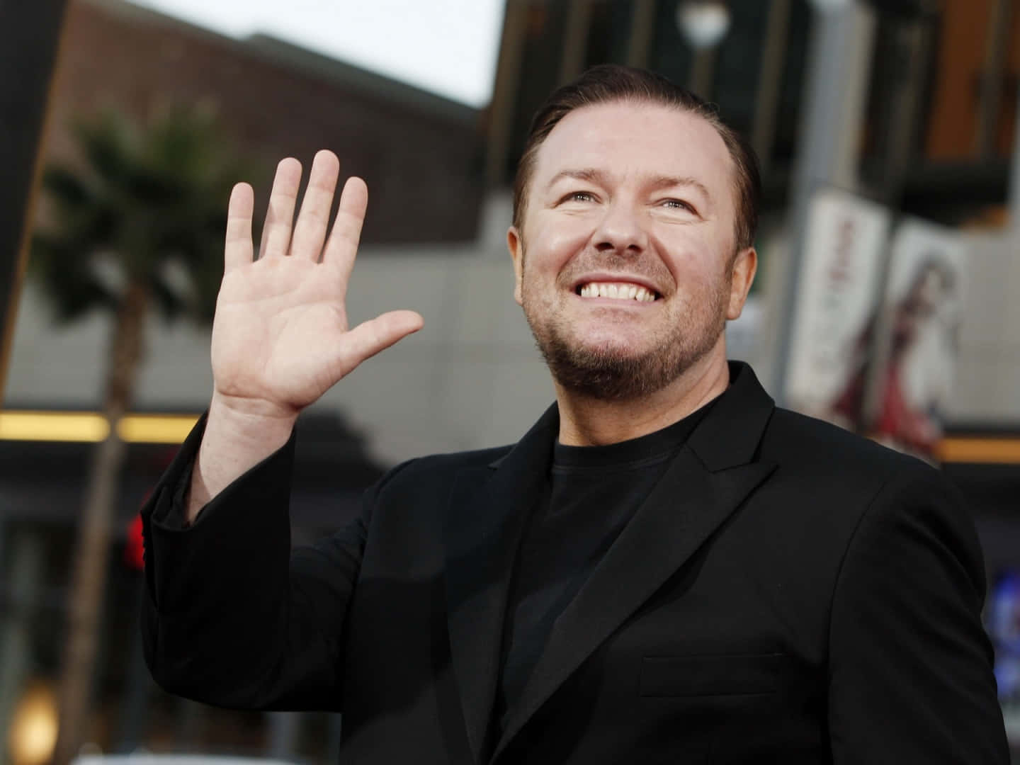 ricky gervais 桌布 壁紙 — 一位身穿黑色西裝的微笑男子，在城市街道上，背景有棕櫚樹和建築物，愉快地向鏡頭揮手。