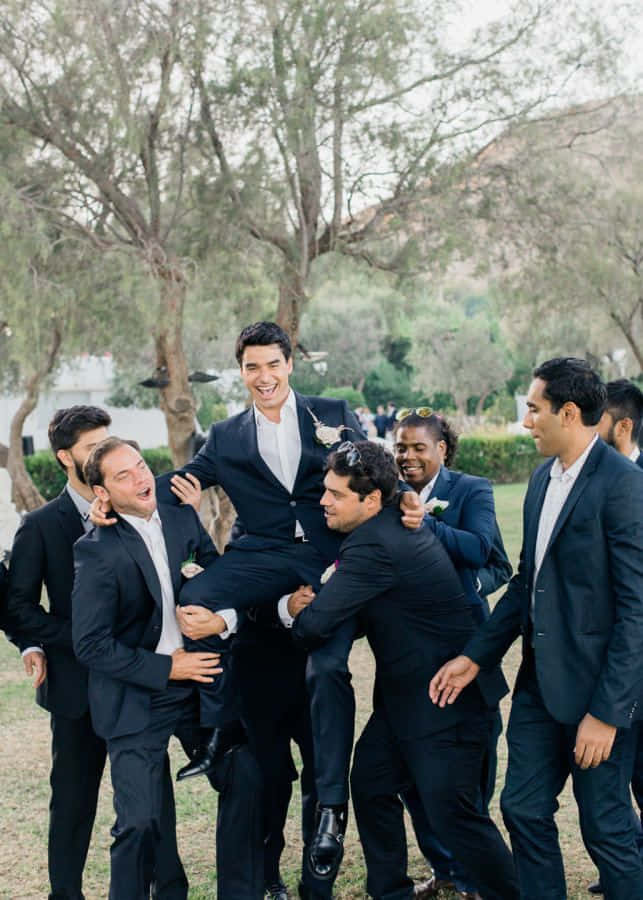 Groomsmen 桌布 壁紙 — 一位身穿黑色西裝的快樂新郎，在枝葉茂密的樹下，被穿著正式服裝、大笑的伴郎們頑皮地抬起。