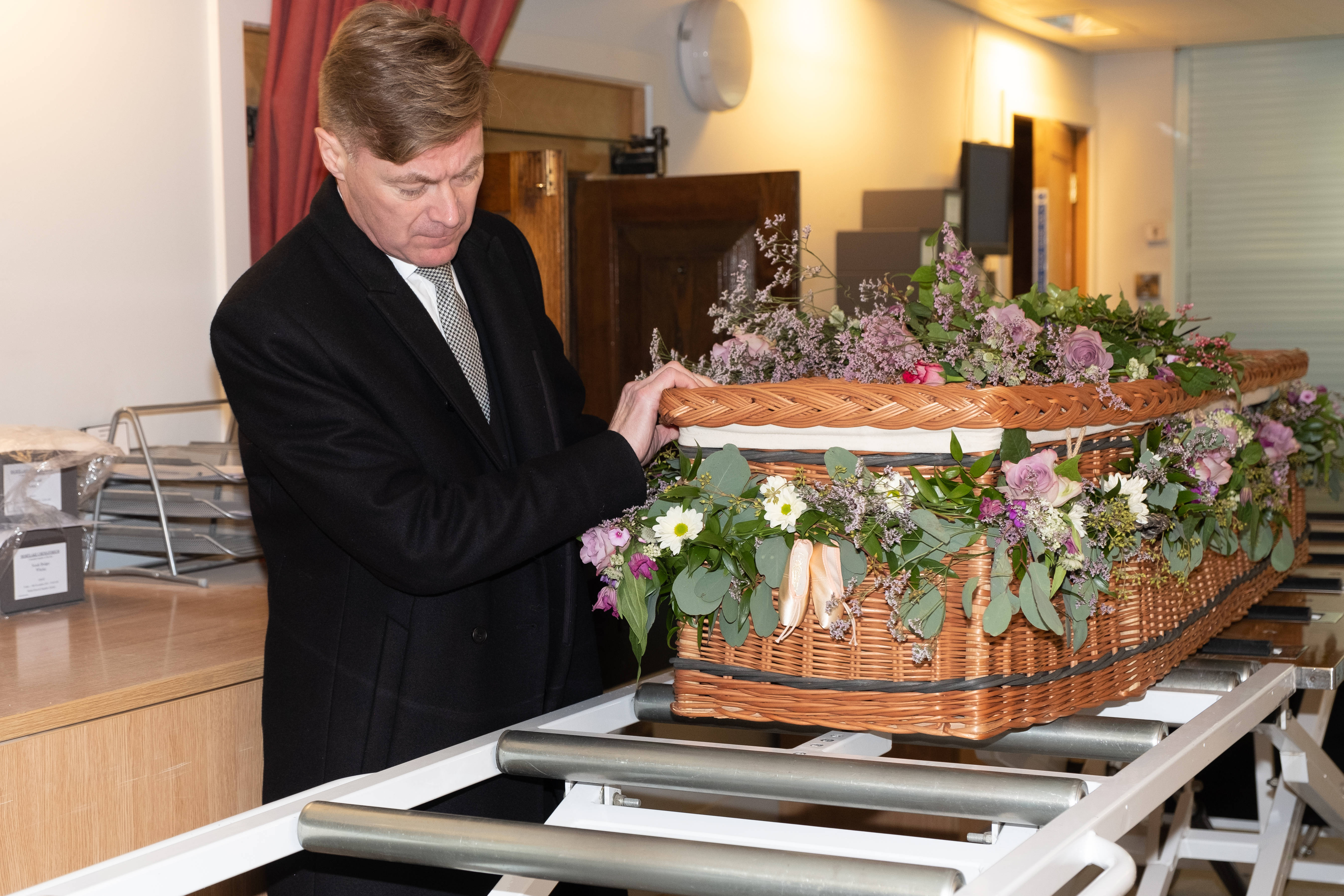Funeral 桌布 壁紙 — 一位身穿黑色西裝的莊嚴男子在殯儀館調整一個飾有紫色、白色和綠色花朵的柳條棺材。