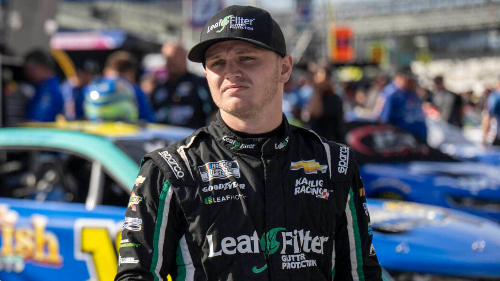 人物 桌布 壁紙 — 一位身穿黑色賽車服和帽子的NASCAR車手在明亮的日光下自信地站在一輛帶有贊助商標誌的藍色賽車旁。
