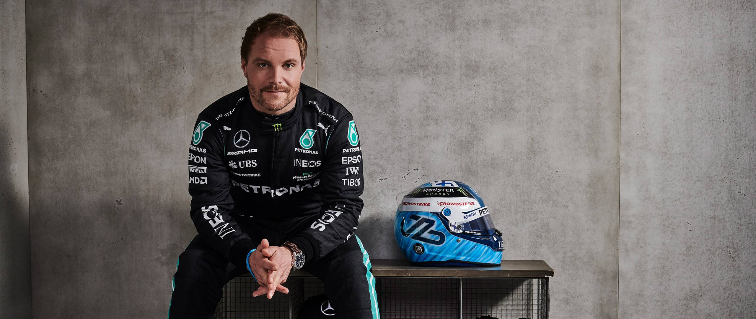 Valtteri Bottas 桌布 壁紙 — 一位身穿黑色賽車服的F1車手，在紋理混凝土牆旁，冷靜地坐在他的藍白頭盔旁邊。