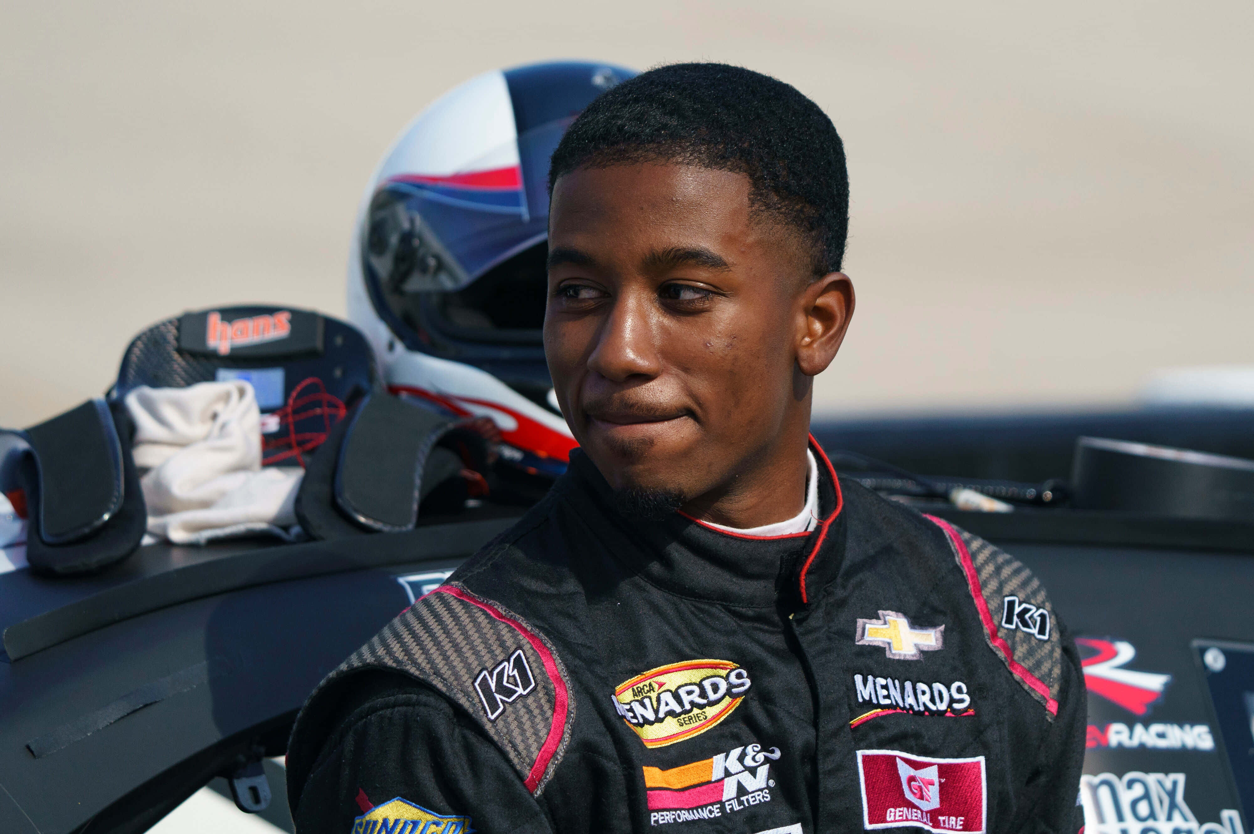 nascar 桌布 壁紙 — 一位身穿黑色贊助商覆蓋賽車服的年輕賽車手站在他的賽車旁邊，頭盔和手套就在附近。