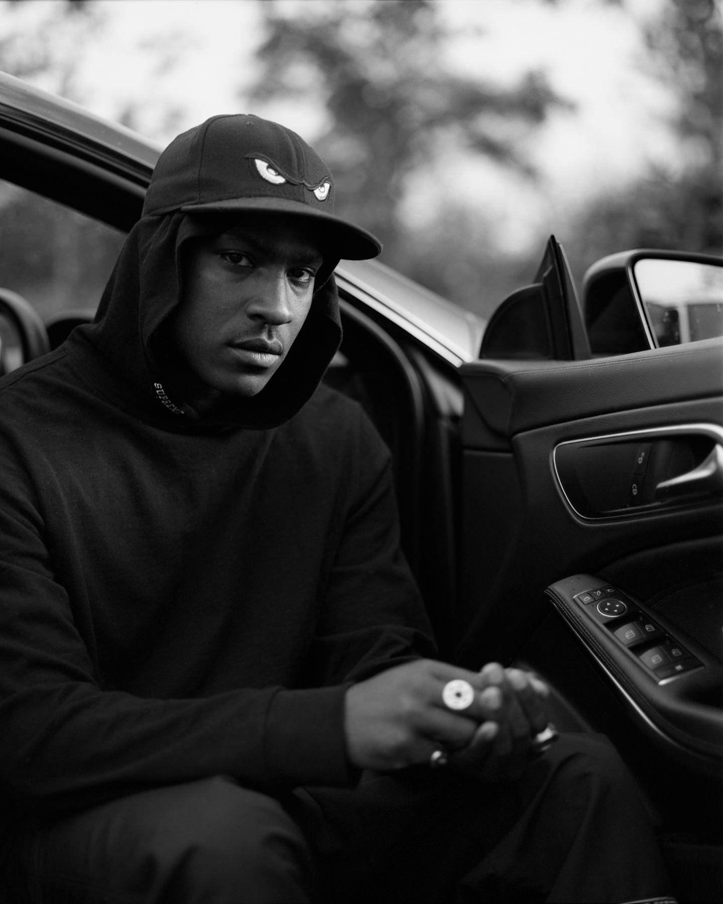 Skepta 桌布 壁紙 — 一位身穿黑色連帽衫和帽子的年輕男子坐在豪華車內，手持電子煙筆，營造出陰鬱的黑白肖像。