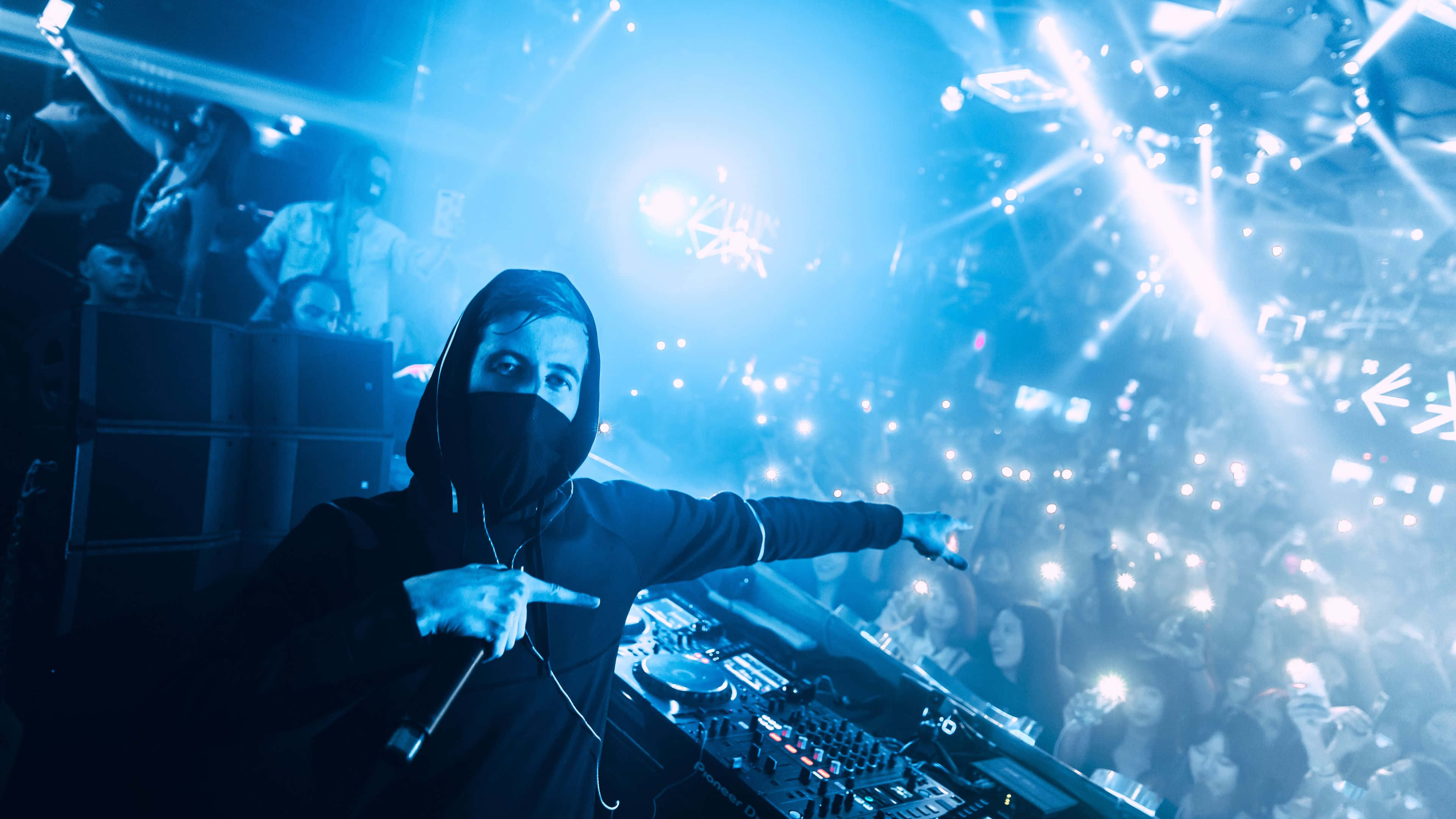 艾倫·沃克 桌布 壁紙 — 一位身穿黑色連帽衫和面具的DJ，舉起雙手，在閃爍的藍色燈光下指揮著夜店的人群。