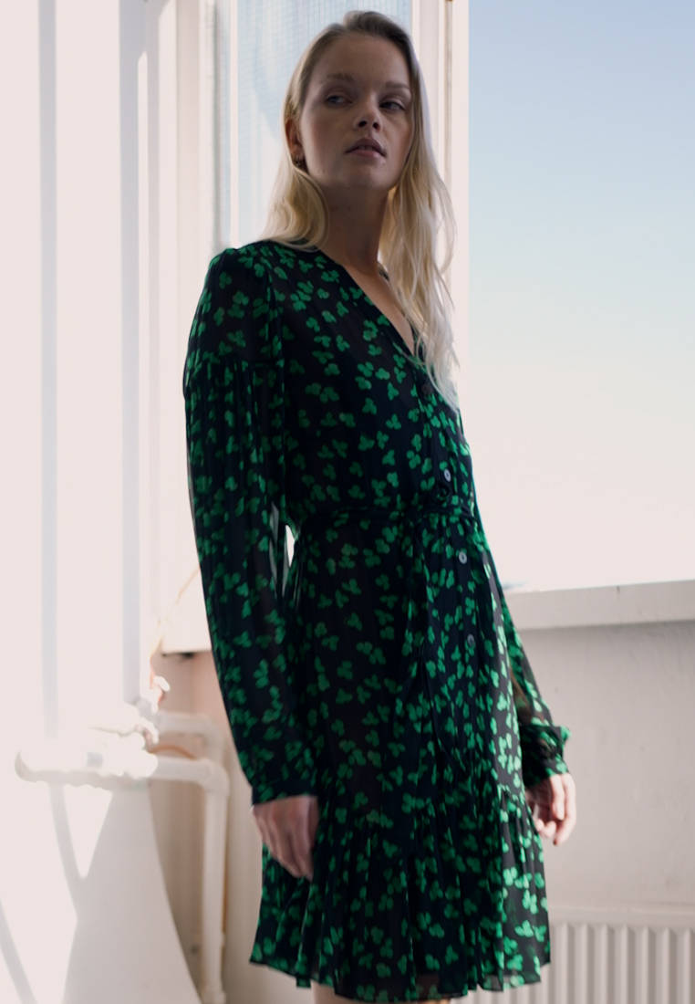 Beautiful Woman 桌布 壁紙 — 一位身穿黑色連衣裙、帶有綠色三葉草圖案的女人站在明亮的窗邊，沐浴在柔和自然光中。