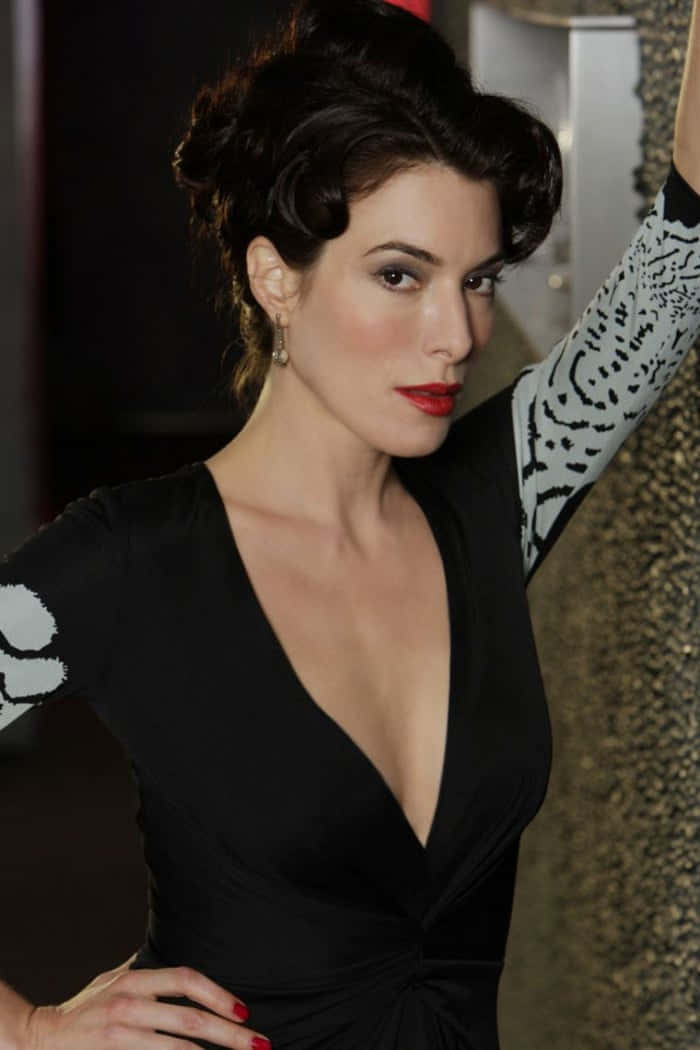 Jaime Murray 桌布 壁紙 — 一位身穿黑色連衣裙、帶白色圖案袖子的優雅女士，自信地在有紋理的牆前擺姿勢。