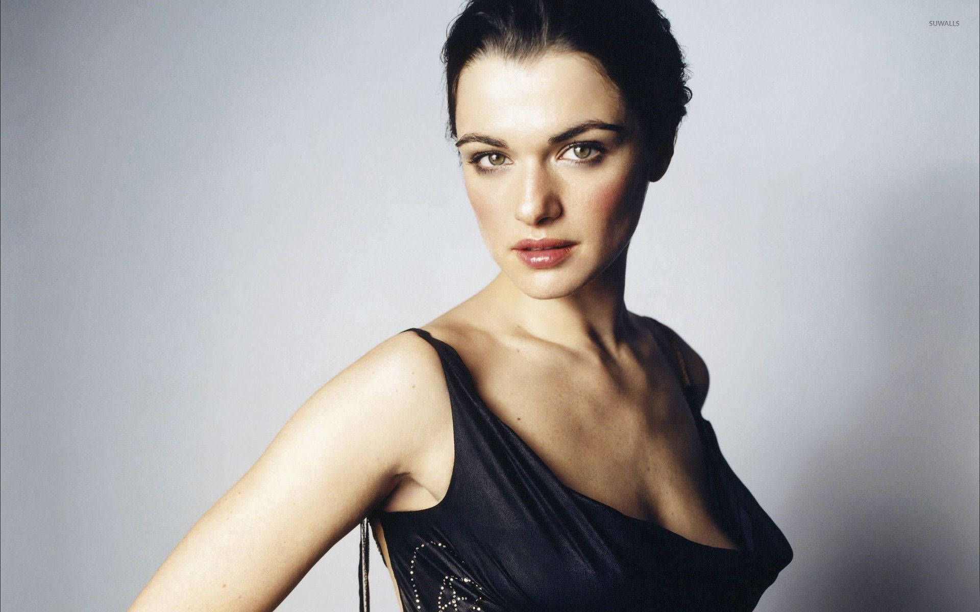 Rachel Weisz 桌布 壁紙 — 一位身穿黑色連衣裙的優雅女士，在柔和的灰色工作室背景下直視鏡頭。