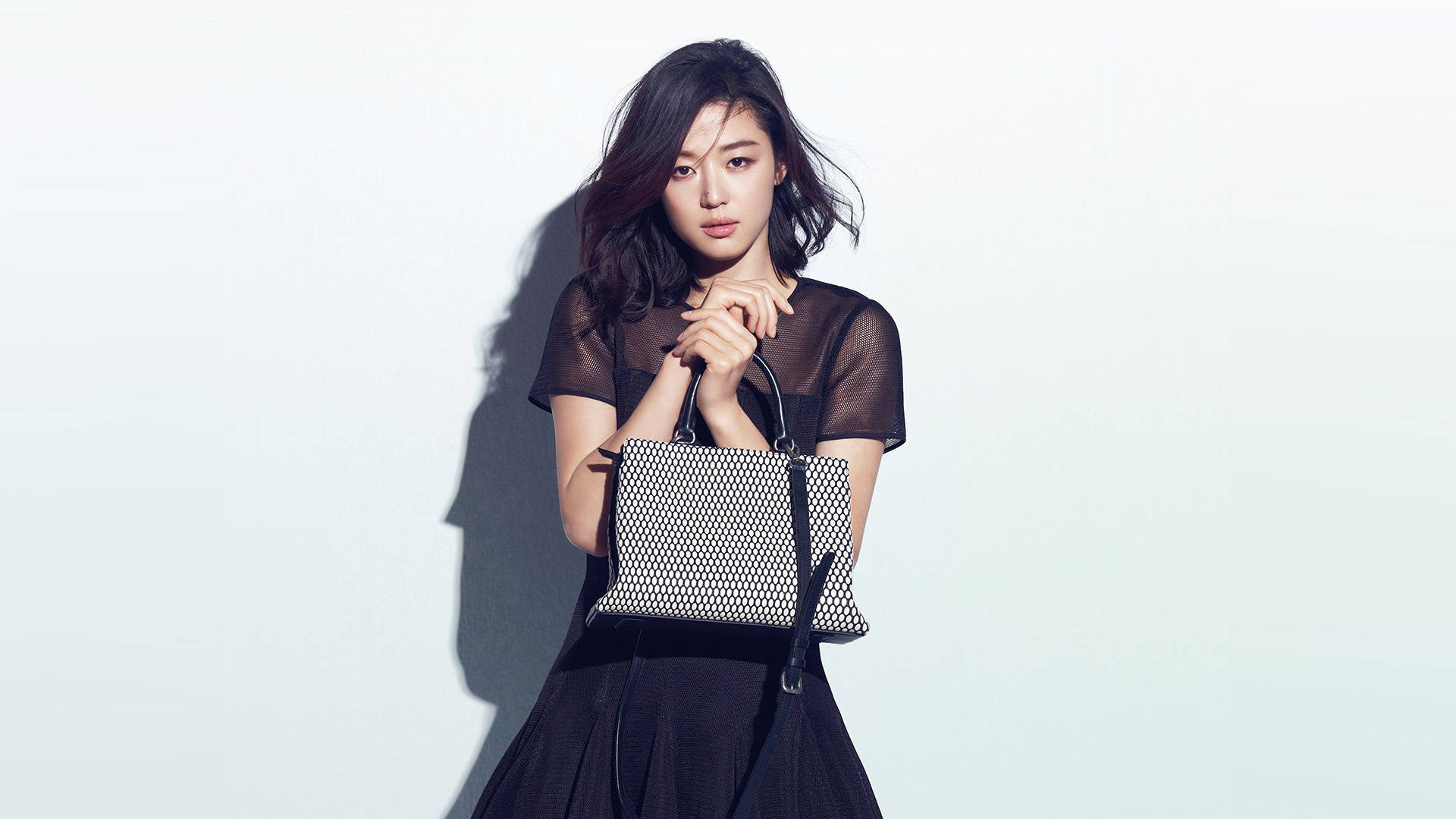 Jun Ji Hyun 桌布 壁紙 — 一位身穿黑色連衣裙的優雅女士，在極簡白色背景上手持圖案手袋，投射出戲劇性的陰影。