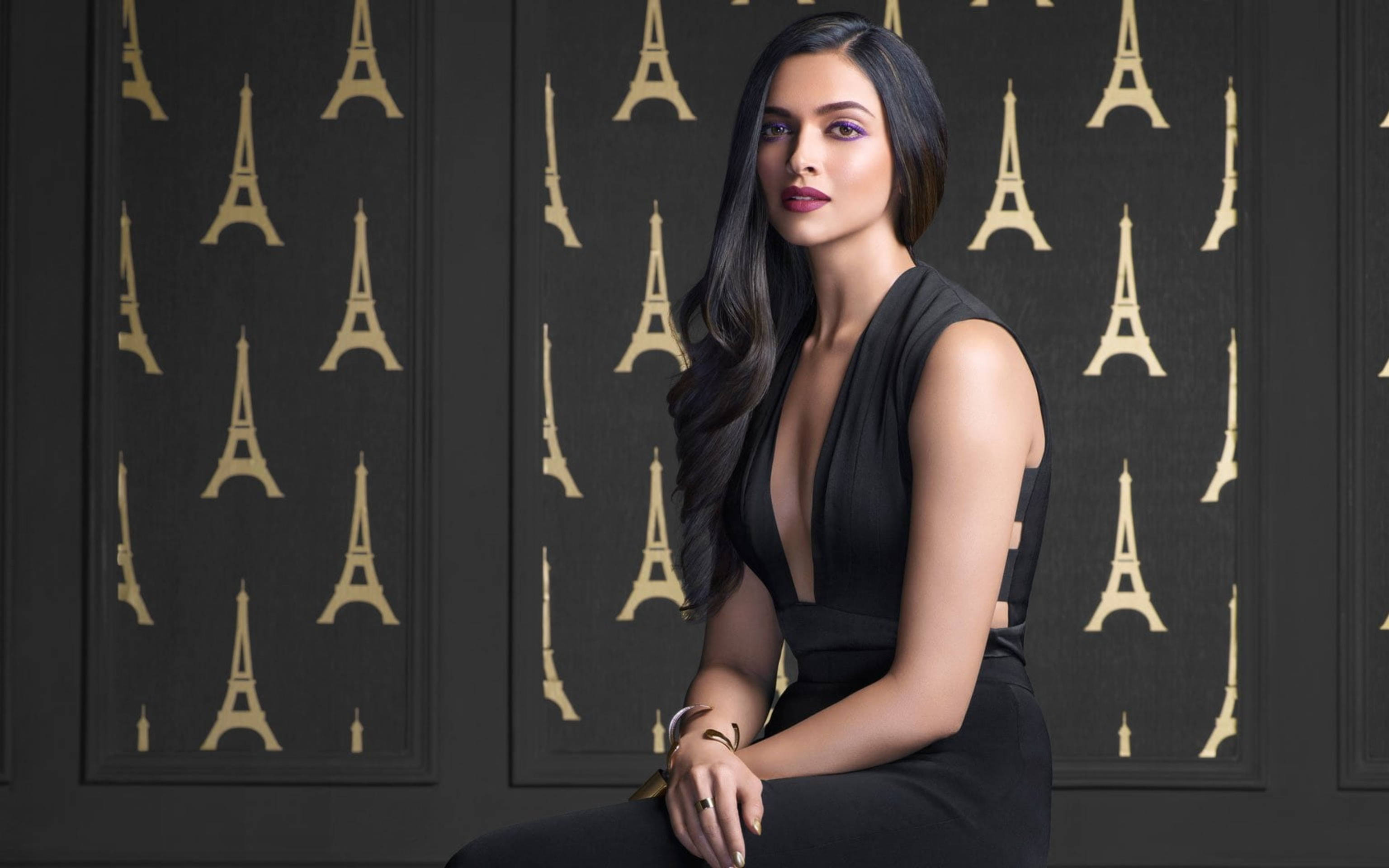 Deepika 桌布 壁紙 — 一位身穿黑色連衣裙的優雅女士，靠著一面飾有重複金色艾菲爾鐵塔圖案的深色牆壁坐著。