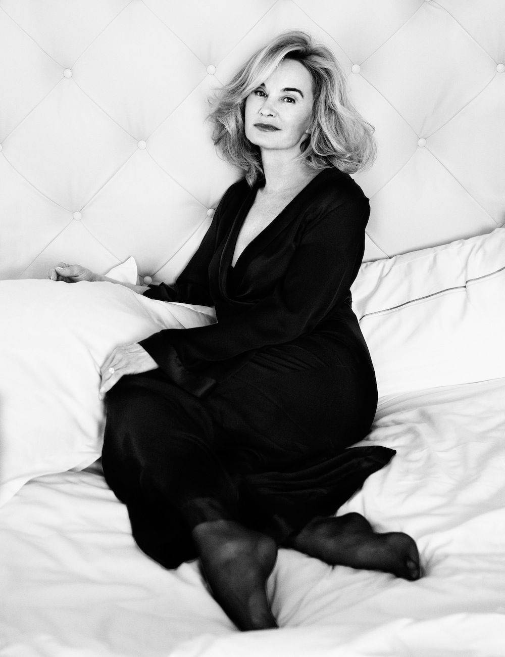 Jessica Lange 桌布 壁紙 — 一位身穿黑色連衣裙的女性優雅地坐在白色簇絨床上，在黑白肖像中若有所思地凝視。