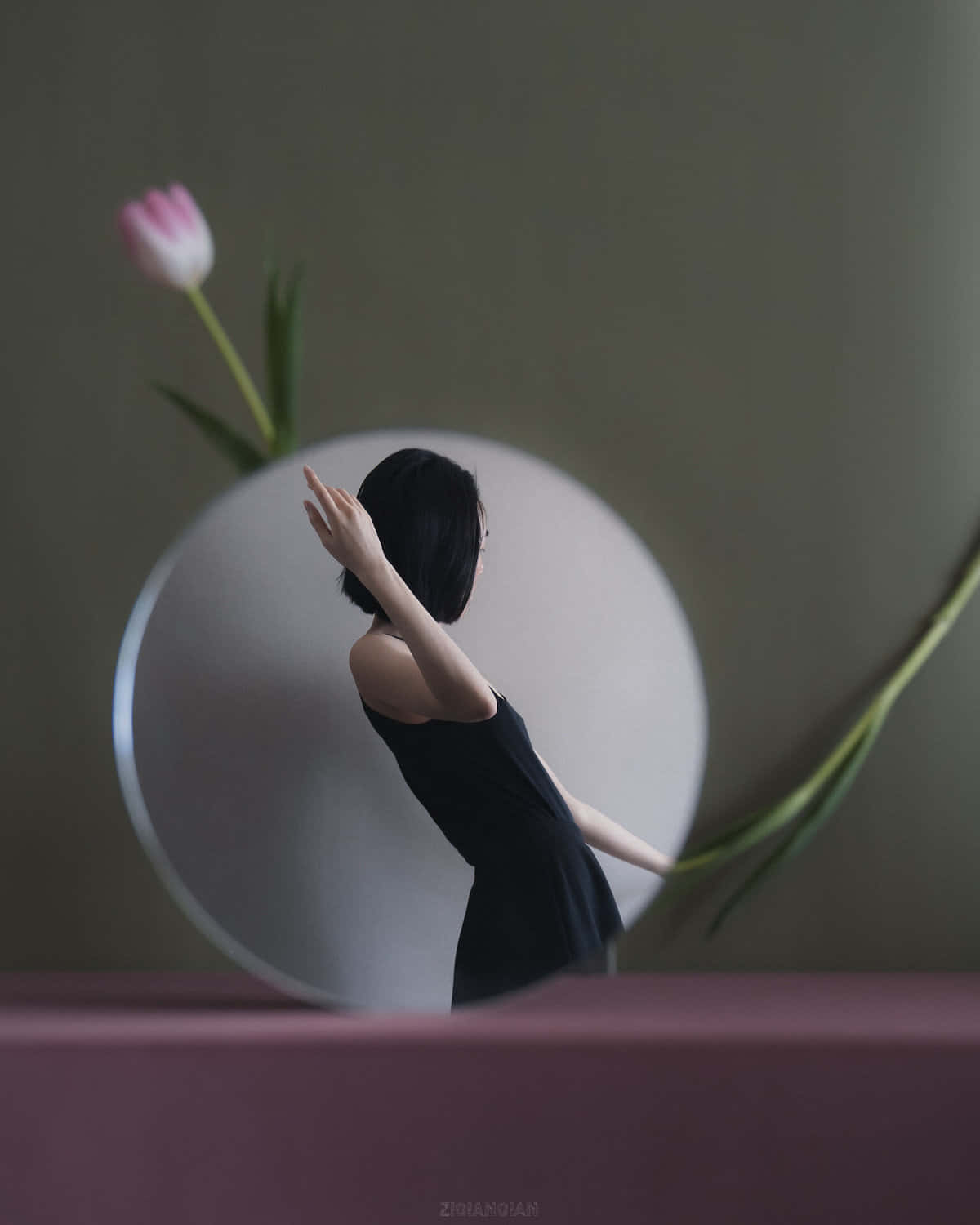 reflection 桌布 壁紙 — 一位身穿黑色連衣裙的女性在一個圓鏡旁邊優雅地伸展雙臂，鏡子旁有一朵柔焦的粉紅色鬱金香和綠色莖。