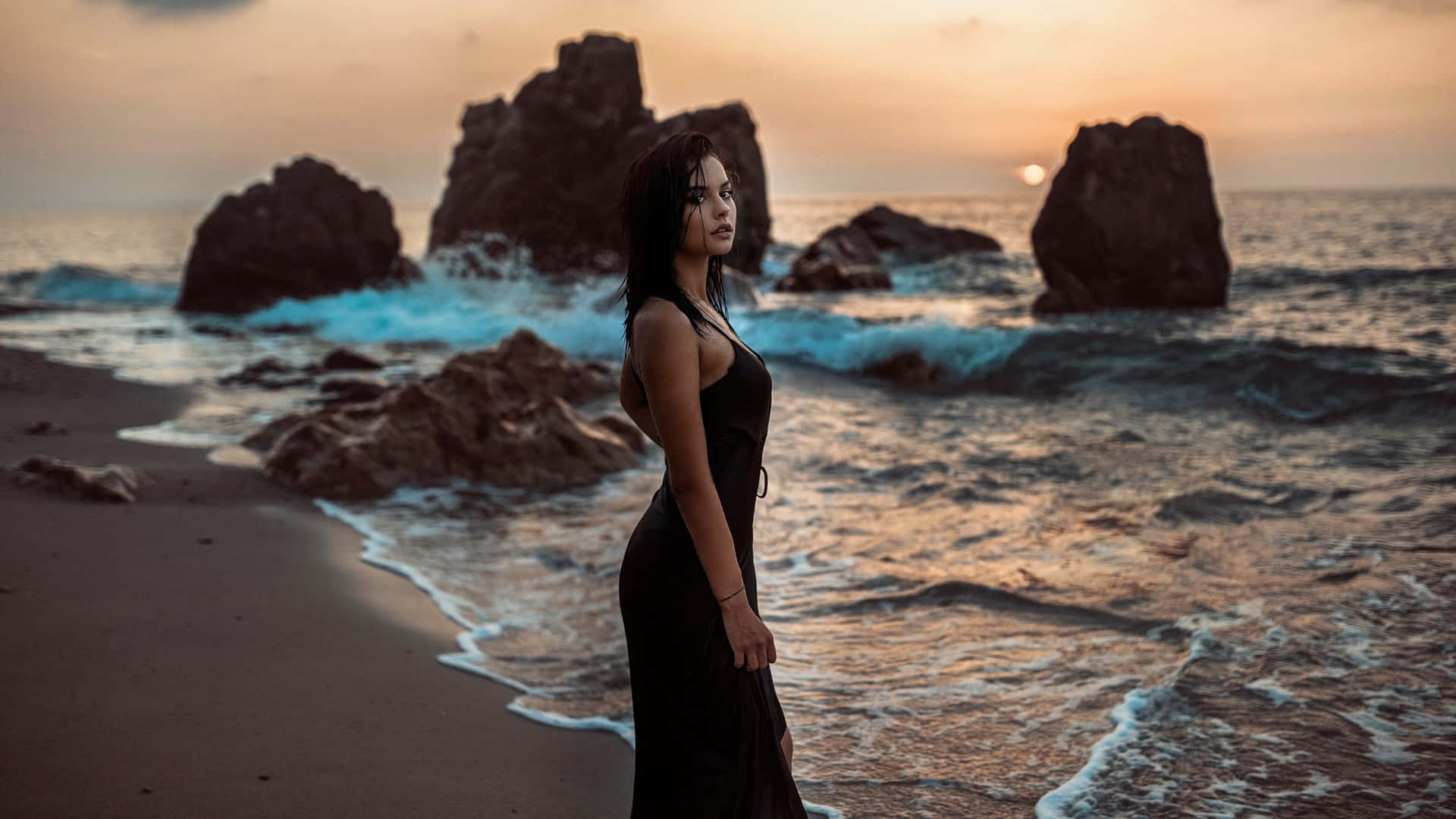 身穿黑色連衣裙的女性在日落時站在海灘上，有海浪和岩石。 — 娜塔莉·波曼 壁紙