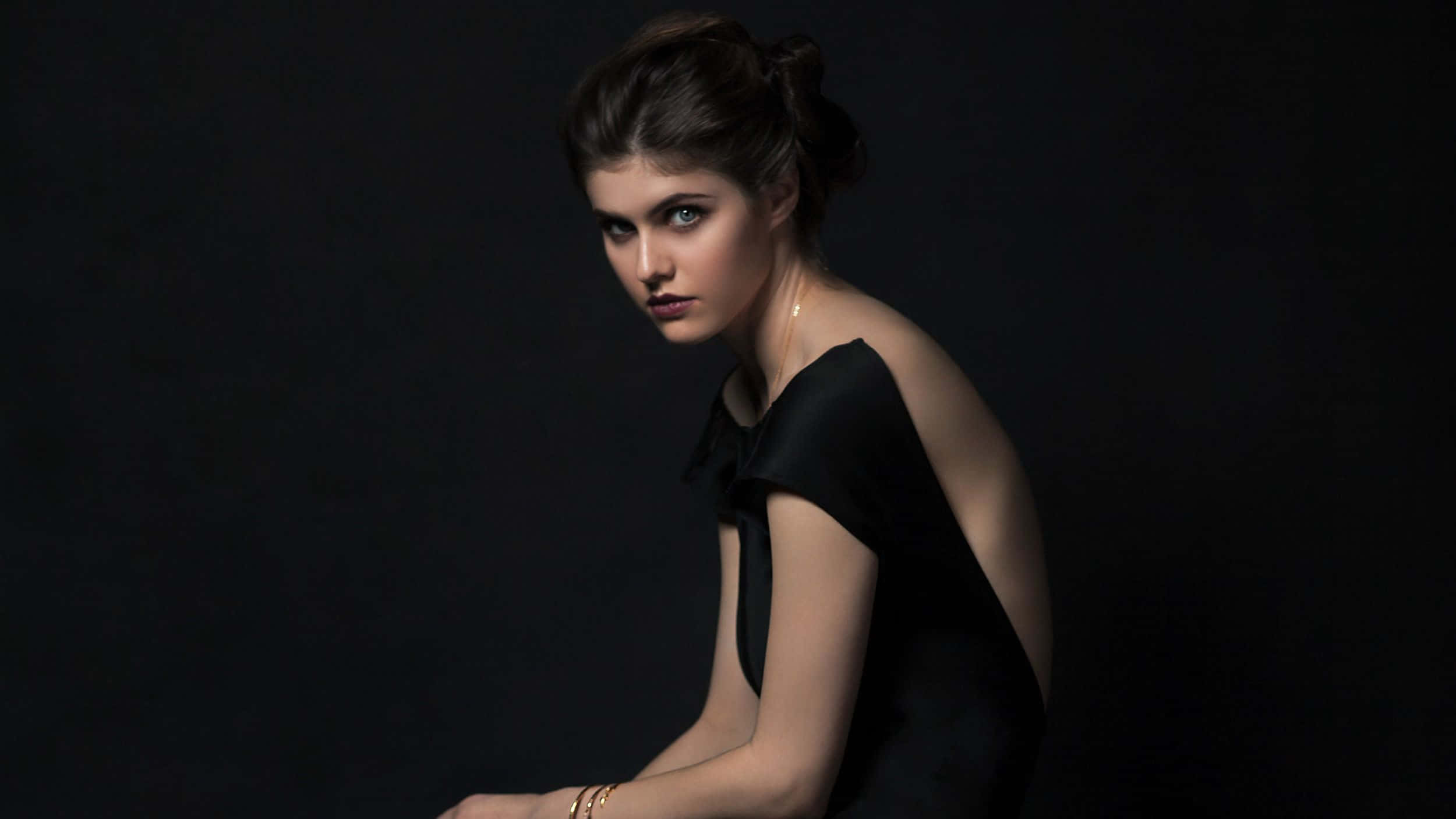 Alexandra Daddario 桌布 壁紙 — 一位身穿黑色連衣裙的女性在黑暗背景下，以戲劇性的燈光突出她的特徵，凝視著前方。