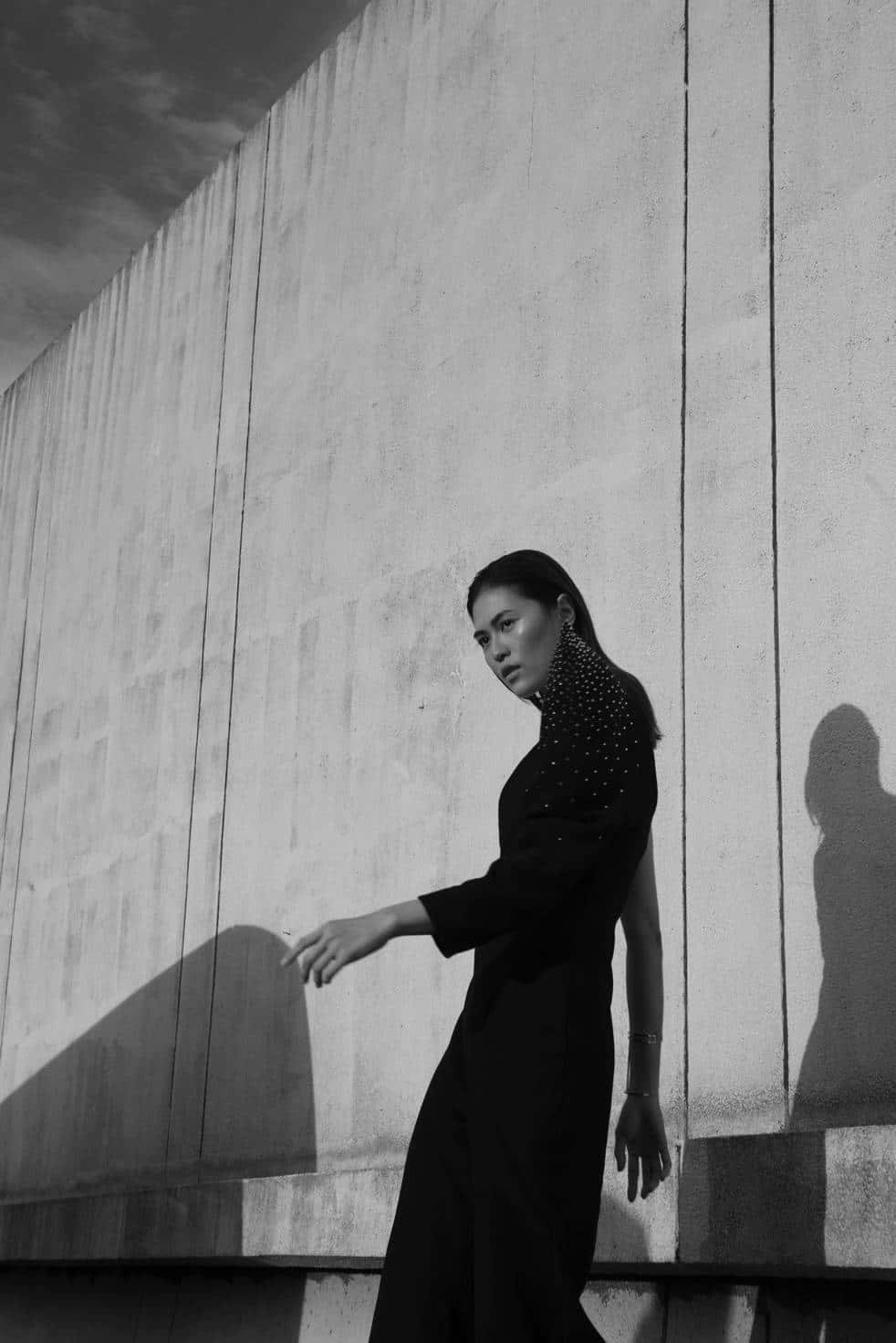 Minimalist Fashion 桌布 壁紙 — 一位身穿黑色連衣裙的女性站在有紋理的混凝土牆前，在單色中投射出戲劇性的陰影。